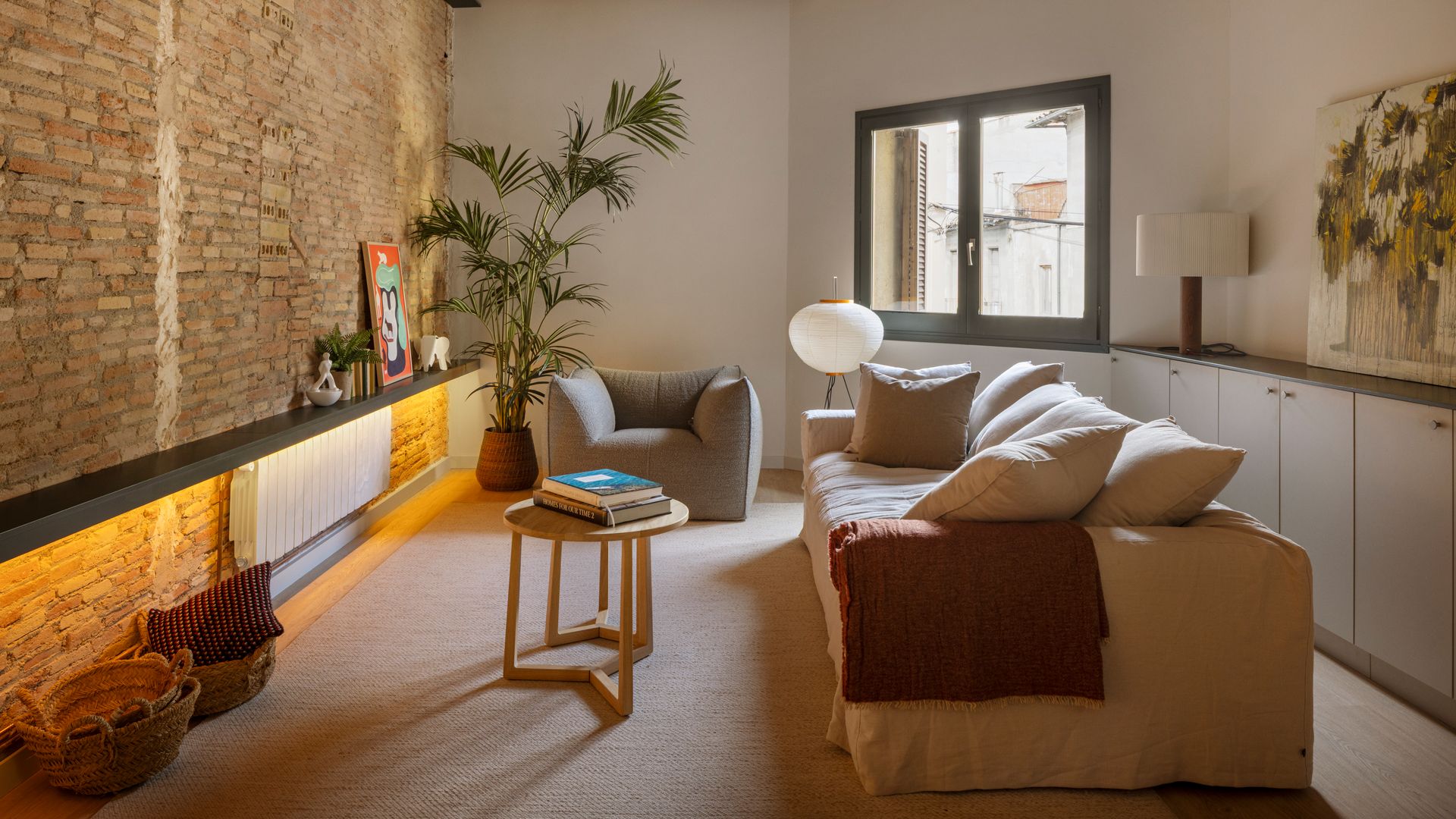 Salón con sofá de blanco con funda, mueble bajo la ventana, pared de ladrillos, alfombra, mesa auxiliar de madera, vigas en el techo, lámpara de pie de papel, sillón gris