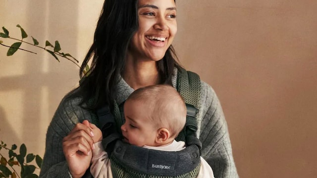 Madre sonriente porteando a su bebé en una mochila de Babybjörn.