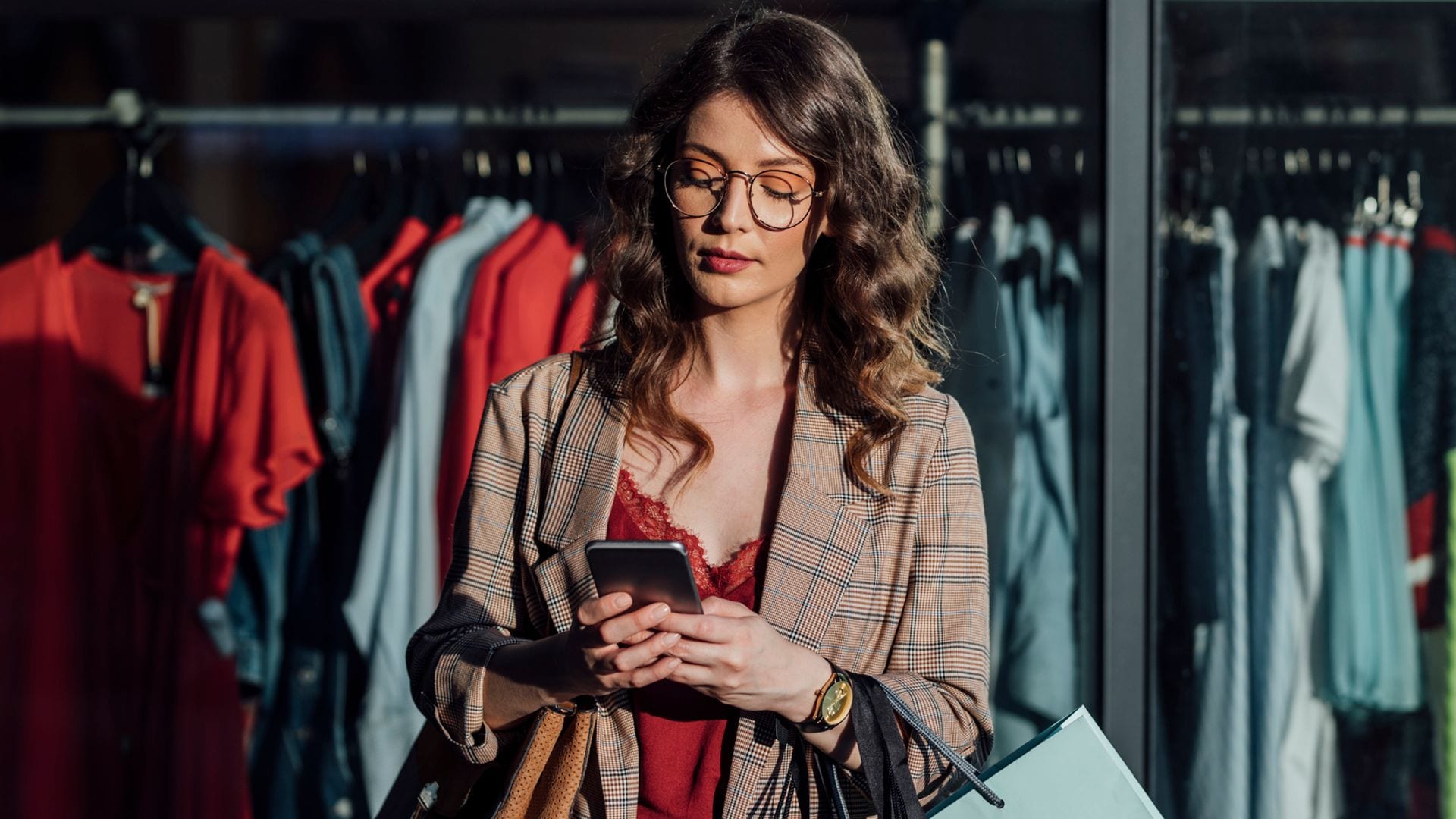 Mujer joven con pelo rizado y gafas mirando su móvil con bolsas de la compra