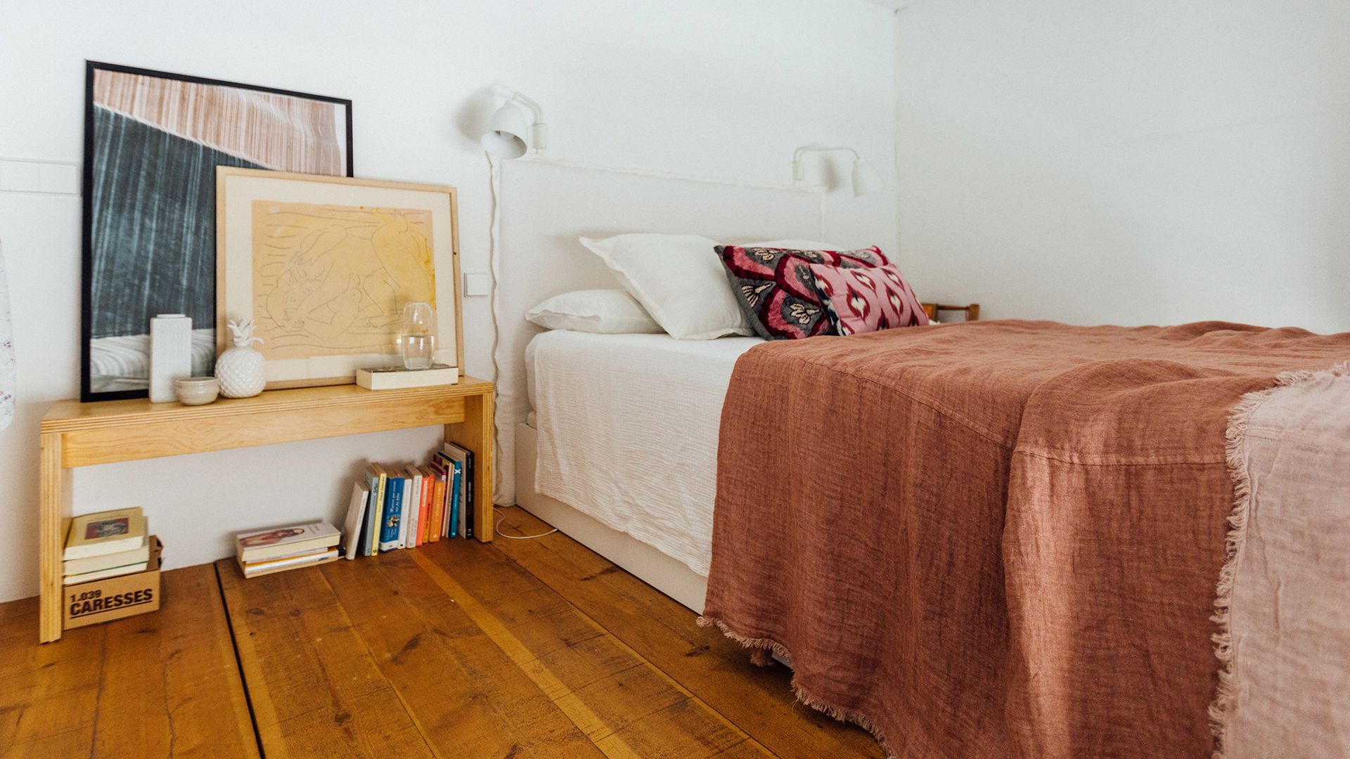 Dormitorio con cabecero tapizado blanco, suelo de madera, banco de madera con cuadros, apliques de pared, plaid marrón, cojines 