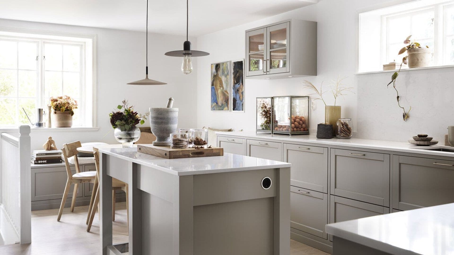 Cocina con isla, muebles grises, mesa de comedor, banco, mueble vitrina, lámparas de techo, ventanas y plantas