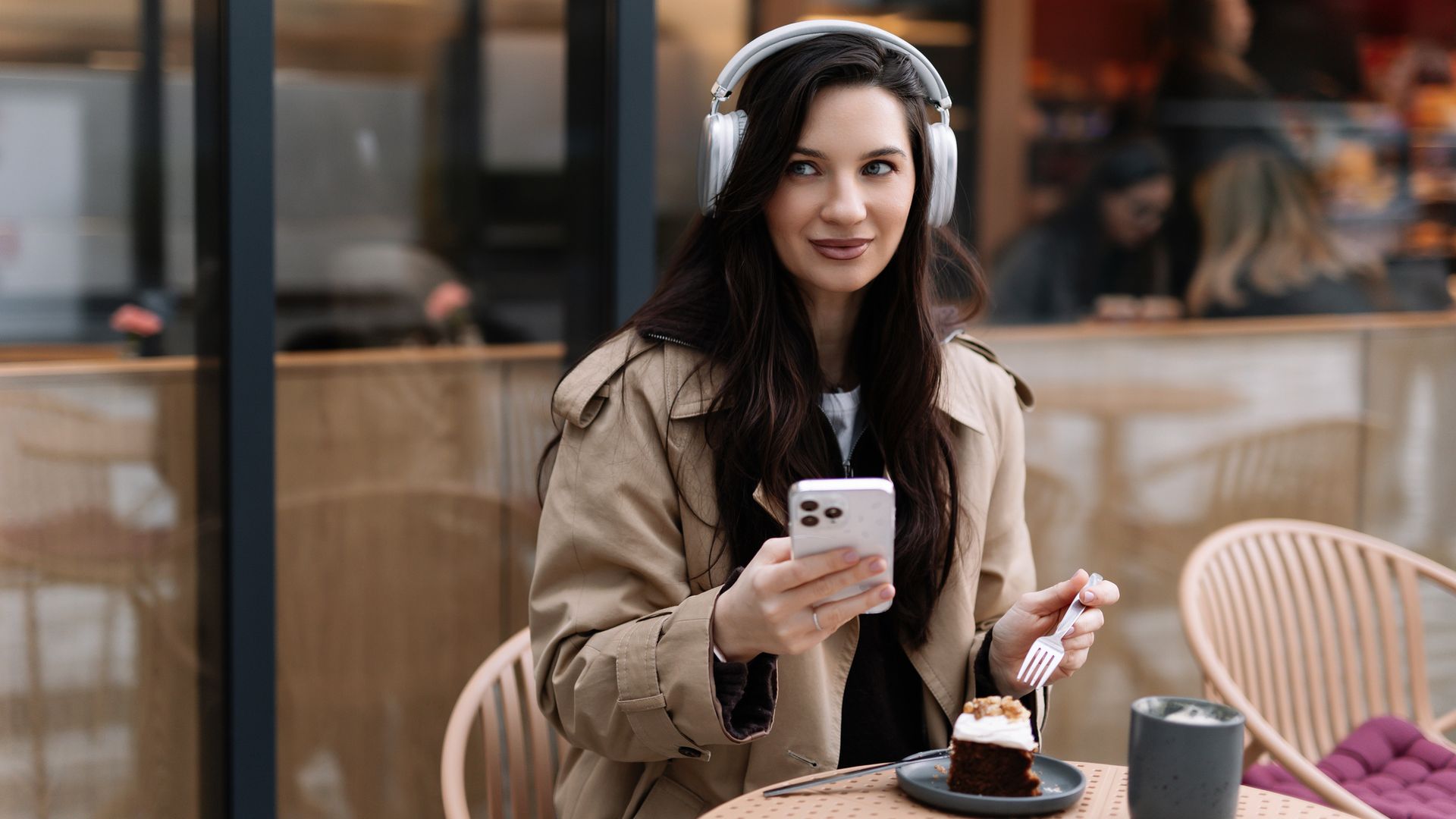 Chica joven de la generación 'millenial' llevando un 'outfit' de otoño con auriculares de diadema, el teléfono en la mano y disfrutando de un café y un trozo de bizcocho en la terraza de un café.
