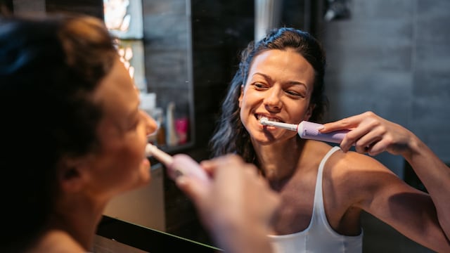 Mujer joven lavándose los dientes con un cepillo eléctrico, mirándose al espejo y sonriendo.