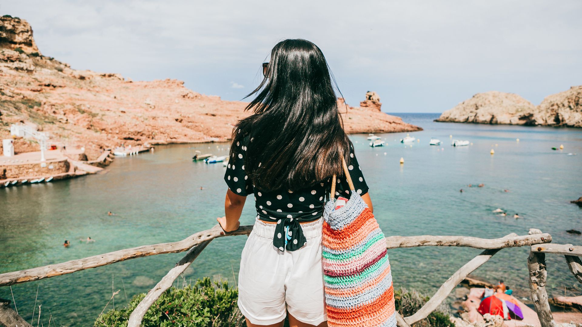 Chica joven asomándose de espaldas a Cala Morell en Menorca (Islas Baleares). Lleva gafas de sol, ropa veraniega y una bolsa de playa de colores.