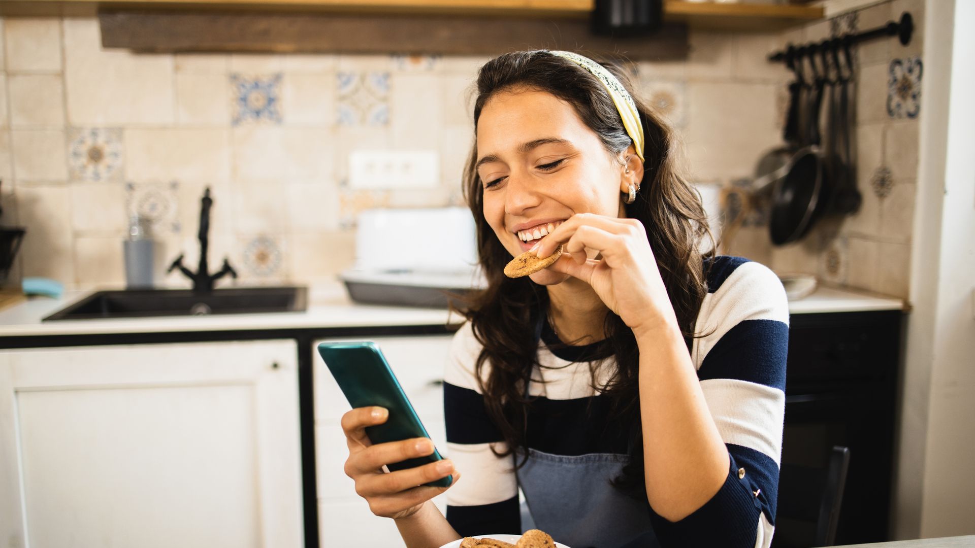 mujer comiendo una galleta en la cocina mientras mira su móvil