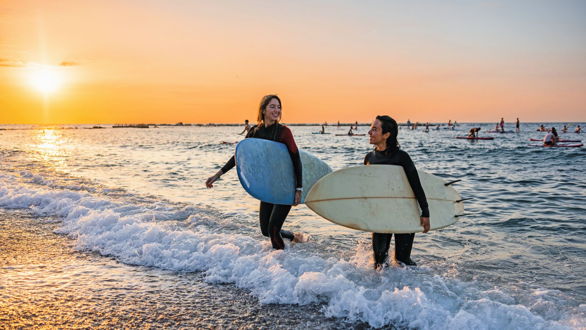 Surferas sonriendo mientras salen del agua de la playa n un atardecer, con tabla de surf y neopreno.