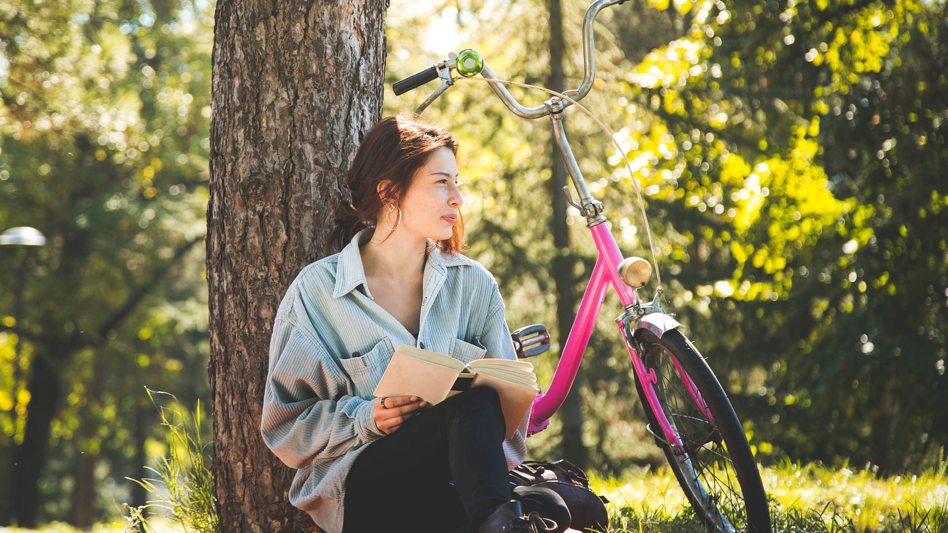 Mujer joven sentada en un parque, apoyada en un árbol con la bici rosa a su lado y leyendo un libro. Lleva ropa de primavera-verano.
