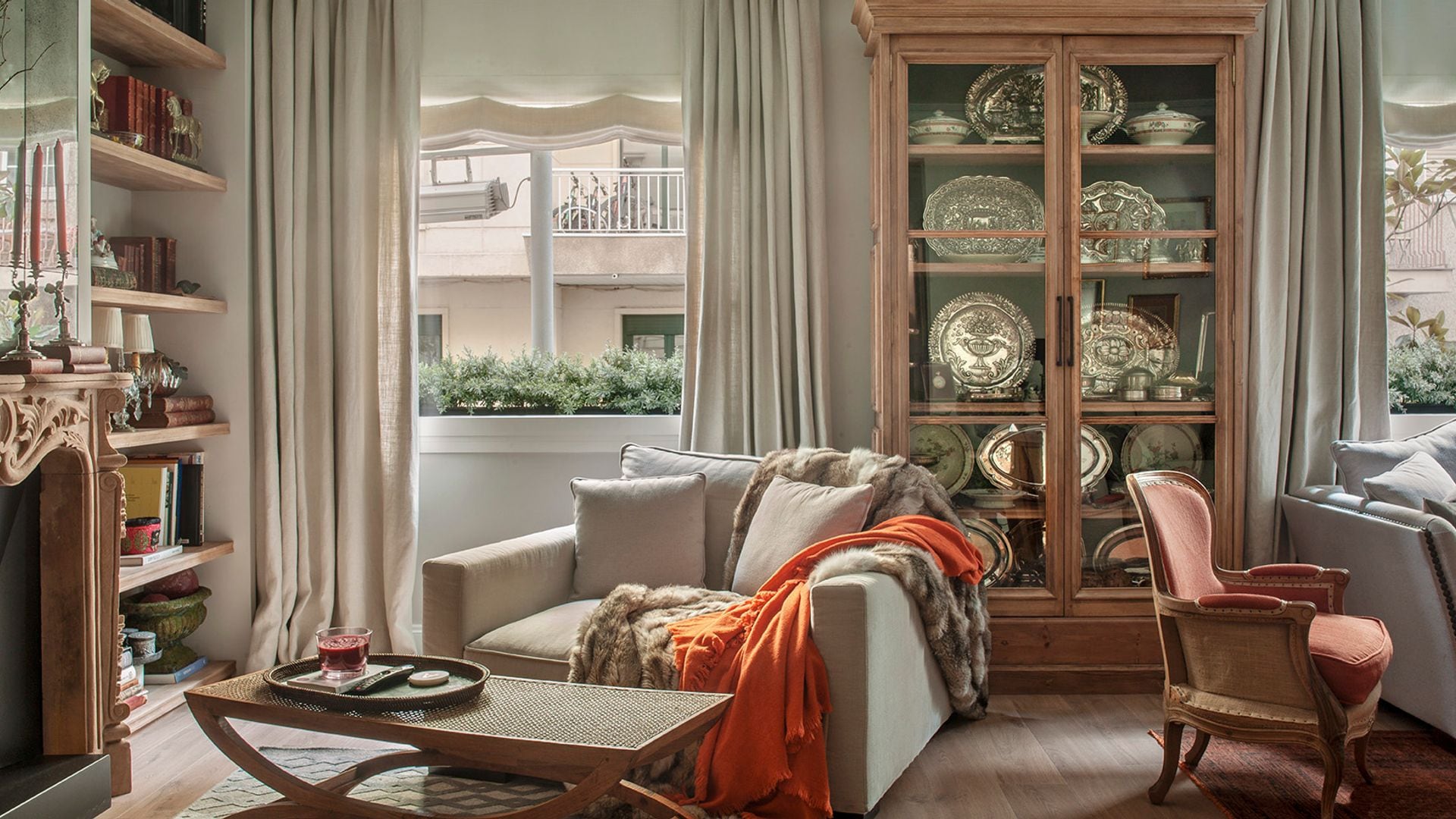 Salón clásico con chimenea, alfombra, sillón, vitrina de madera con piezas de plata, butaca y estantería