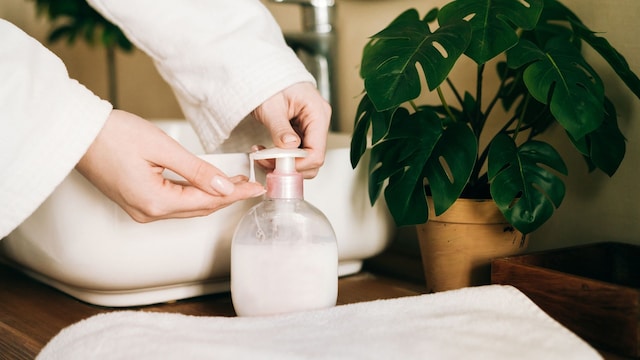 Dispenser with white liquid soap or shower gel, for intimate hygiene, cream. Woman's hands pushing a button. White toilet soap in female hands against the background of a fragrant foam bath. Woman in white terry bathrobe washing her hands with soap. Relaxation and beauty treatments. Natural beauty, daily skincare routine. Moisturizing, cleansing