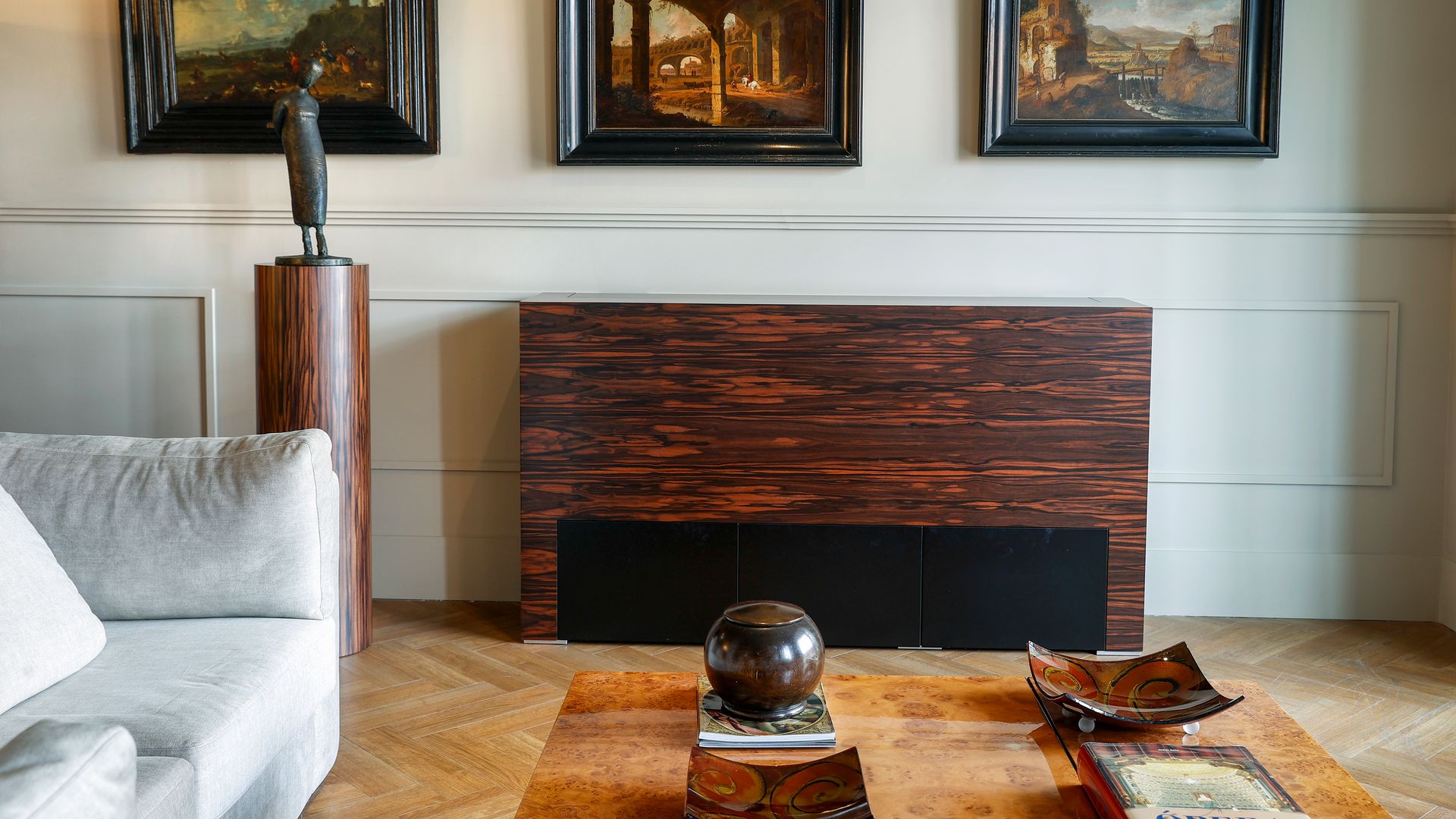 Salón con chimenea de madera oscura, sofá blanco, suelo de madera de espiga, friso de pared, escultura con pedestal, mesa de centro de madera, cuadros en la pared