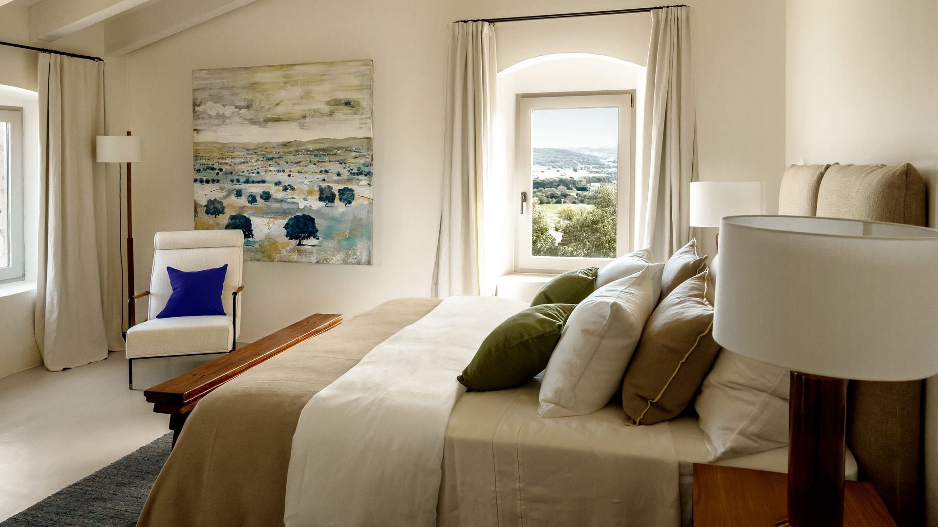 Dormitorio con ventanas, rincón de lectura con cojín azul Klein, cuadro en la pared, cama con cojines crudos, marrones y verdes, lámparas de sobremesa, cabecero tapizado