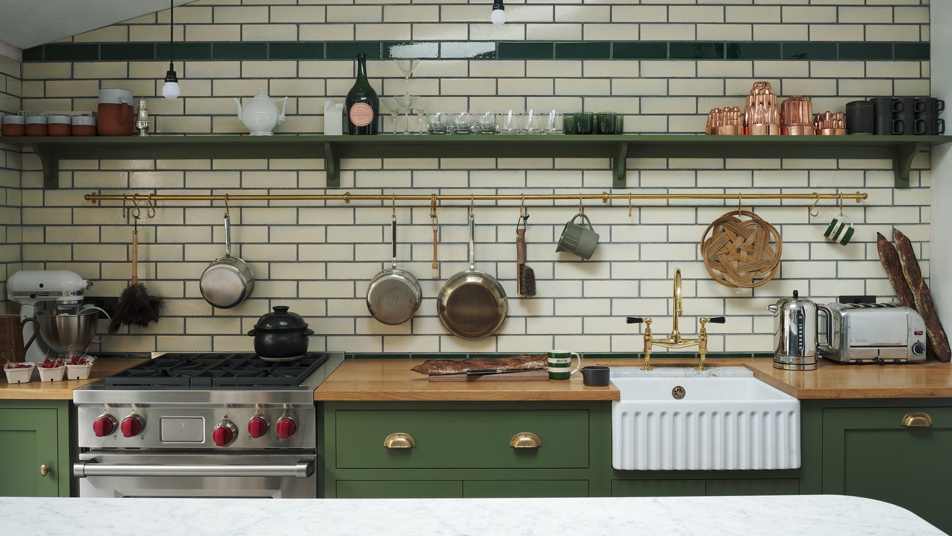 Cocina con muebles verdes, encimera de madera, paredes de azulejos blancos, barra dorada con utensilios y sartenes colgando, balda verde con botes y accesorios 
