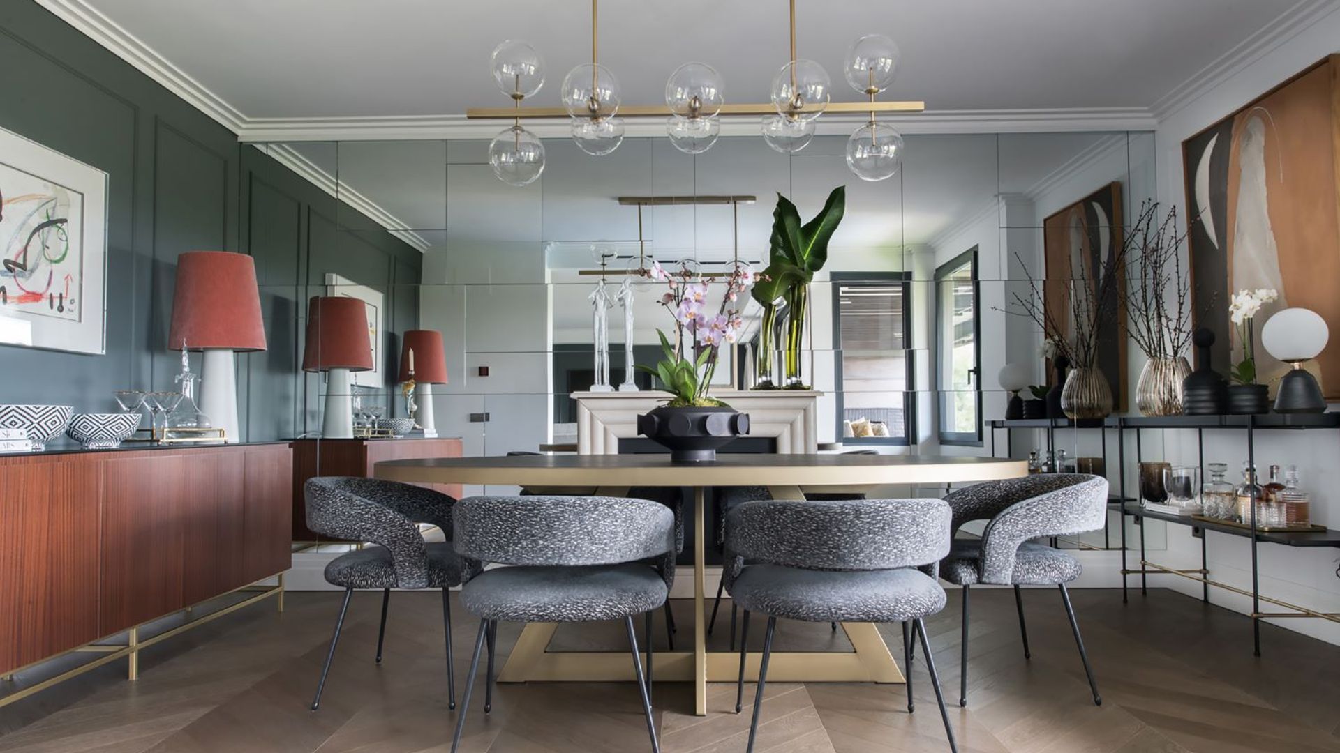 Comedor con sillas tapizadas en gris, pared de espejo, aparador de madera, mesa de comedor, lámpara de techo, cuadros en las paredes, estantería negra 
