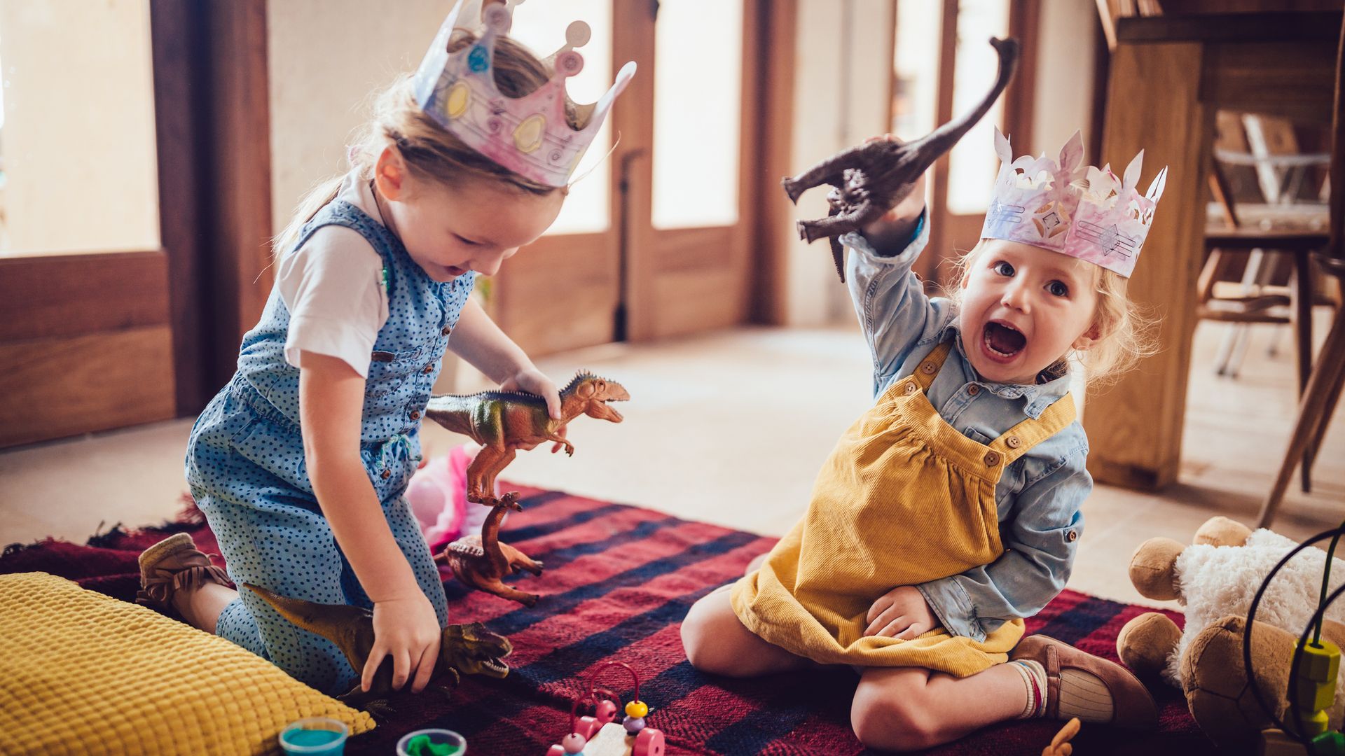 Dos hermanas pequeñas jugando con dinosaurios, plastilinas y otros juguetes, en el suelo sobre una alfombra. Llevan coronas de papel en la cabeza.
