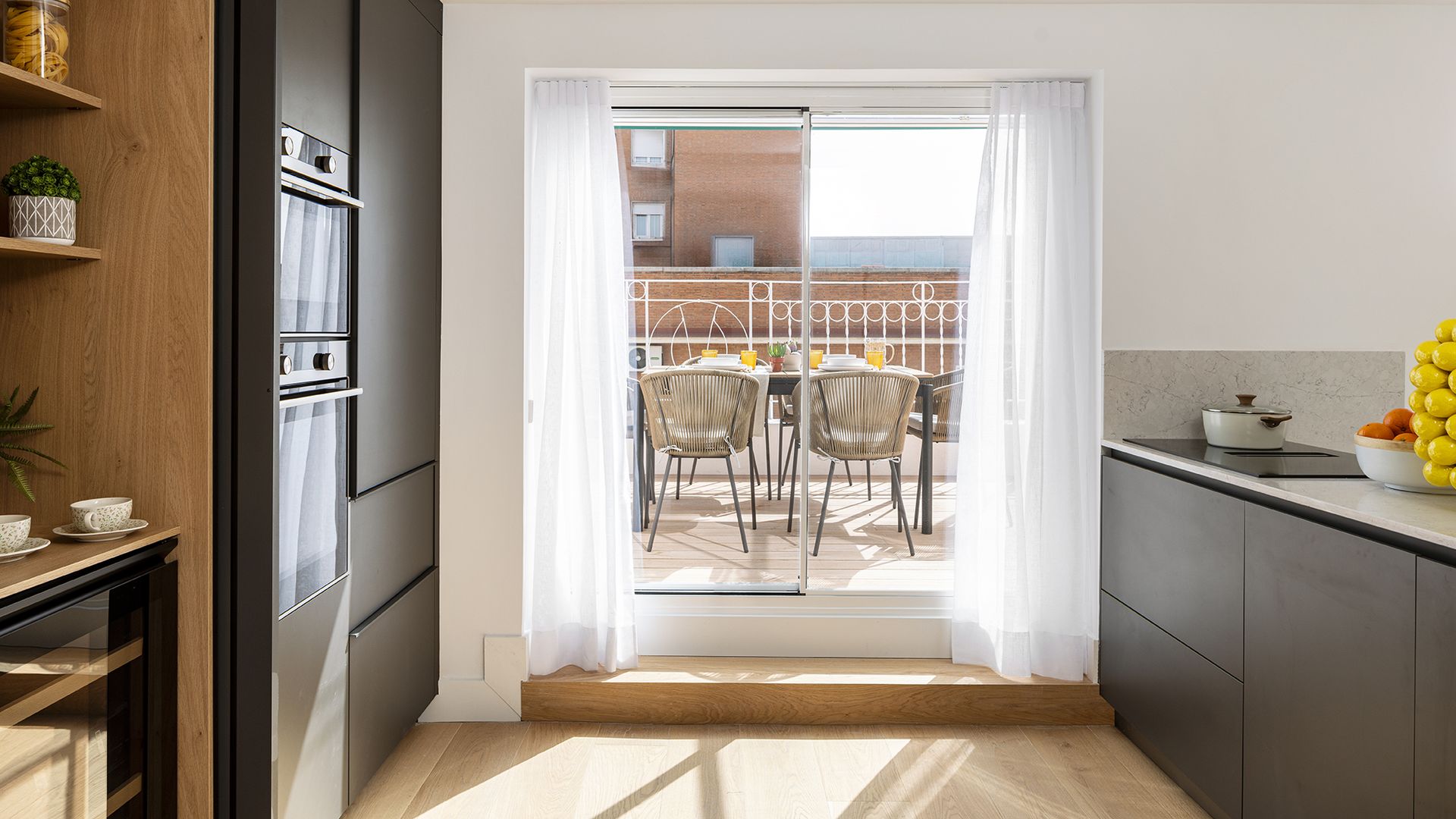 Cocina abierta a la terraza con comedor exterior, cortinas blancas, muebles de cocina gris grafito 