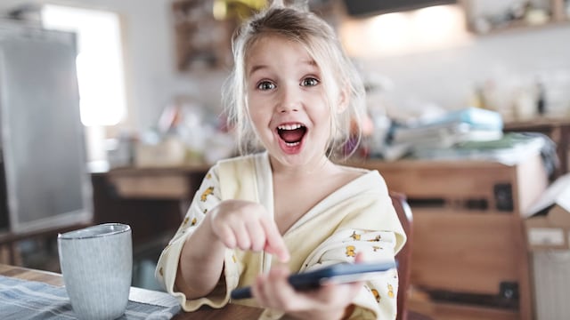Retrato de una niña emocionada en la cocina señalando un teléfono inteligente.