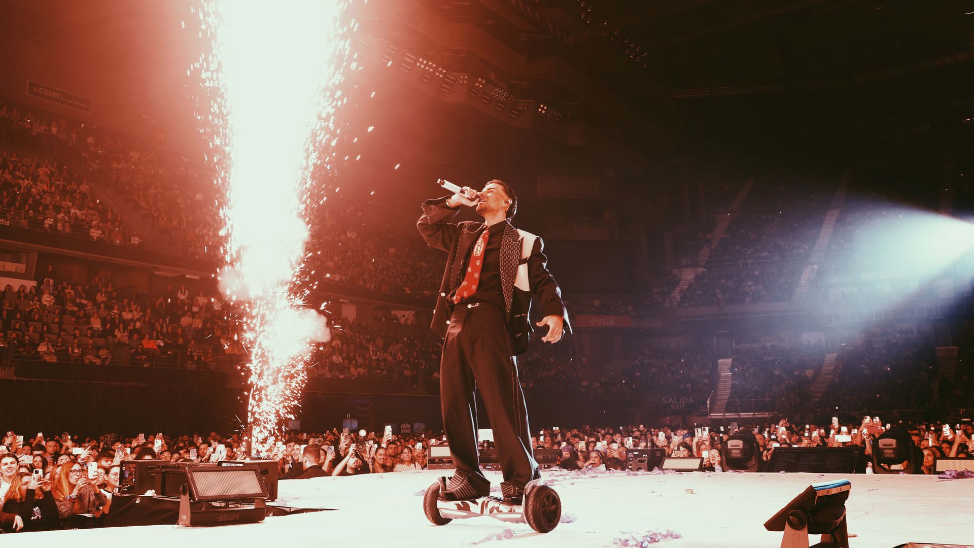 Abraham Mateo en traje y corbata rojos, cantando en un segway en un estadio lleno de gente.