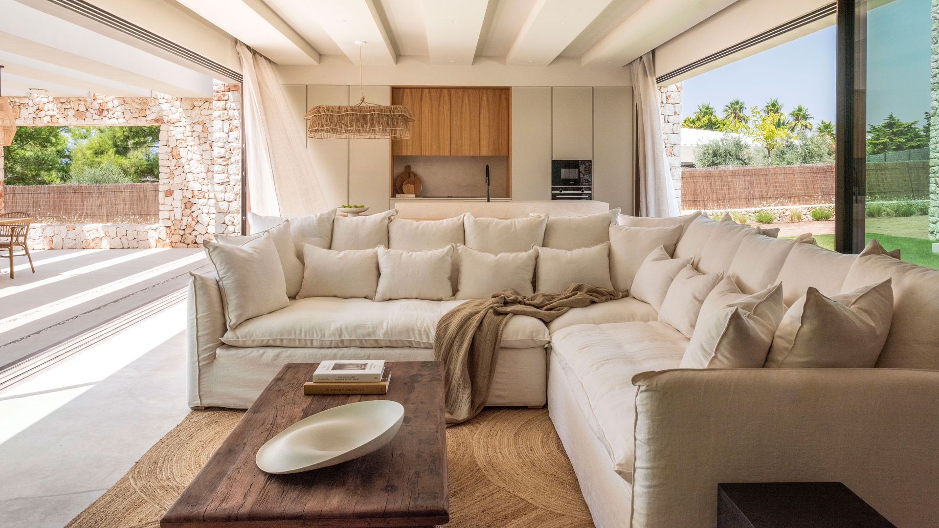 Espacio único con salón y cocina abierto al jardín por dos lado, con sofá de lino beige, alfombra de fibras vegetales, mesa de madera, vigas techo