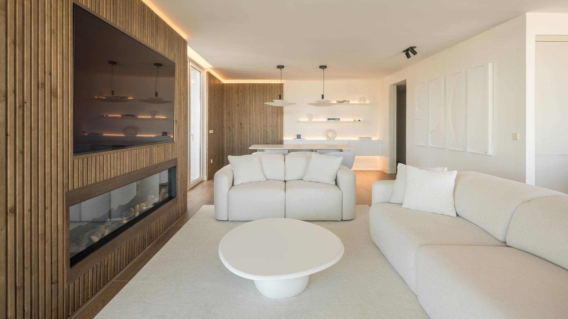 Salón con televisión colgada en pared de madera, sofás blancos, mesa de centro blanca, alfombra blanca, iluminación perimétral en el techo, chimenea moderna