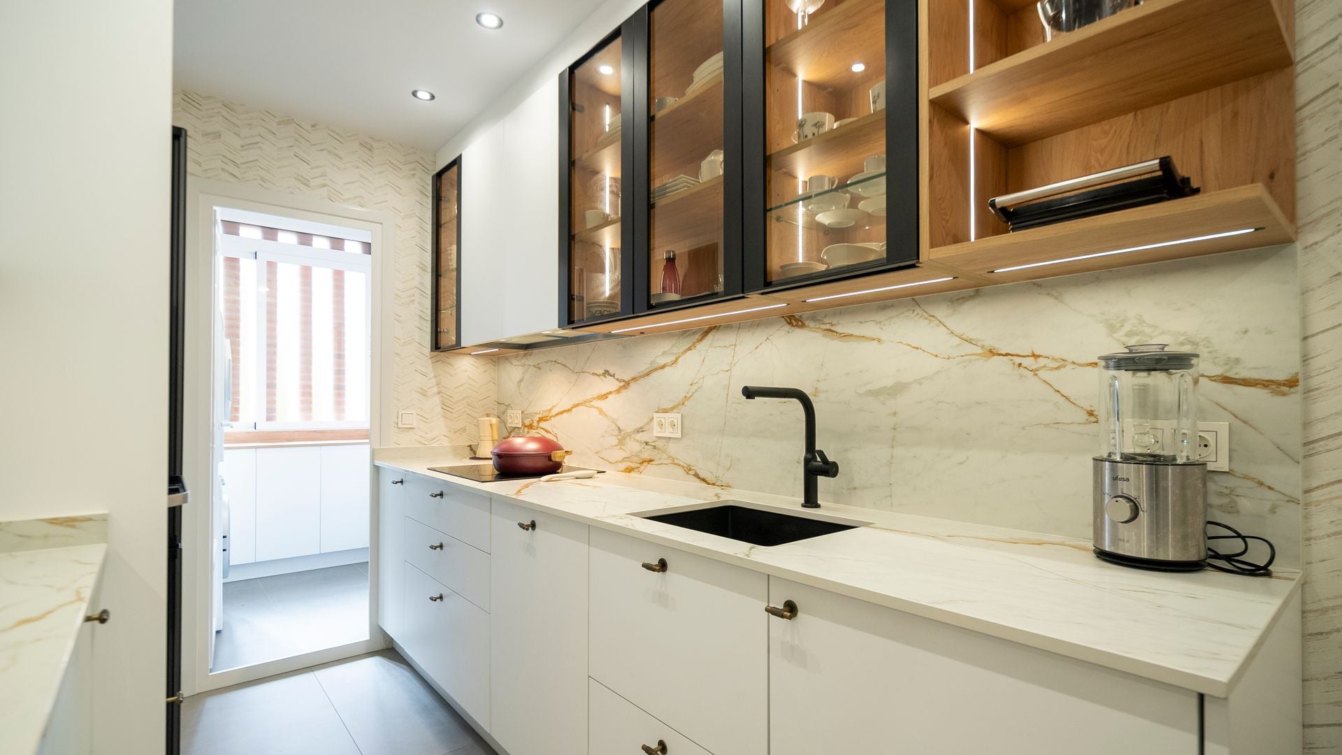 Cocina con muebles y encimera blancos, armarios superiores de vitrina con luz interior, salida tendedero, focos empotrados, grifo negro