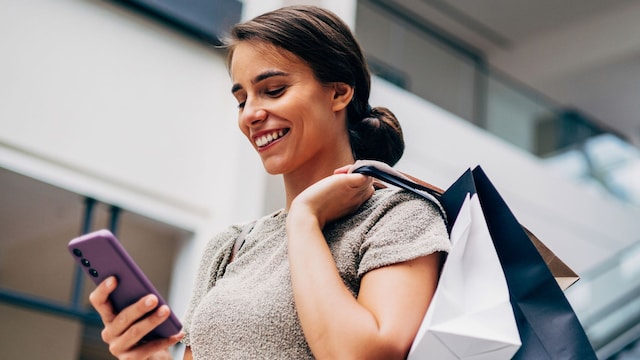 Mujer joven sonriente en centro comercial con bolsas de compras y teléfono móvil en la mano