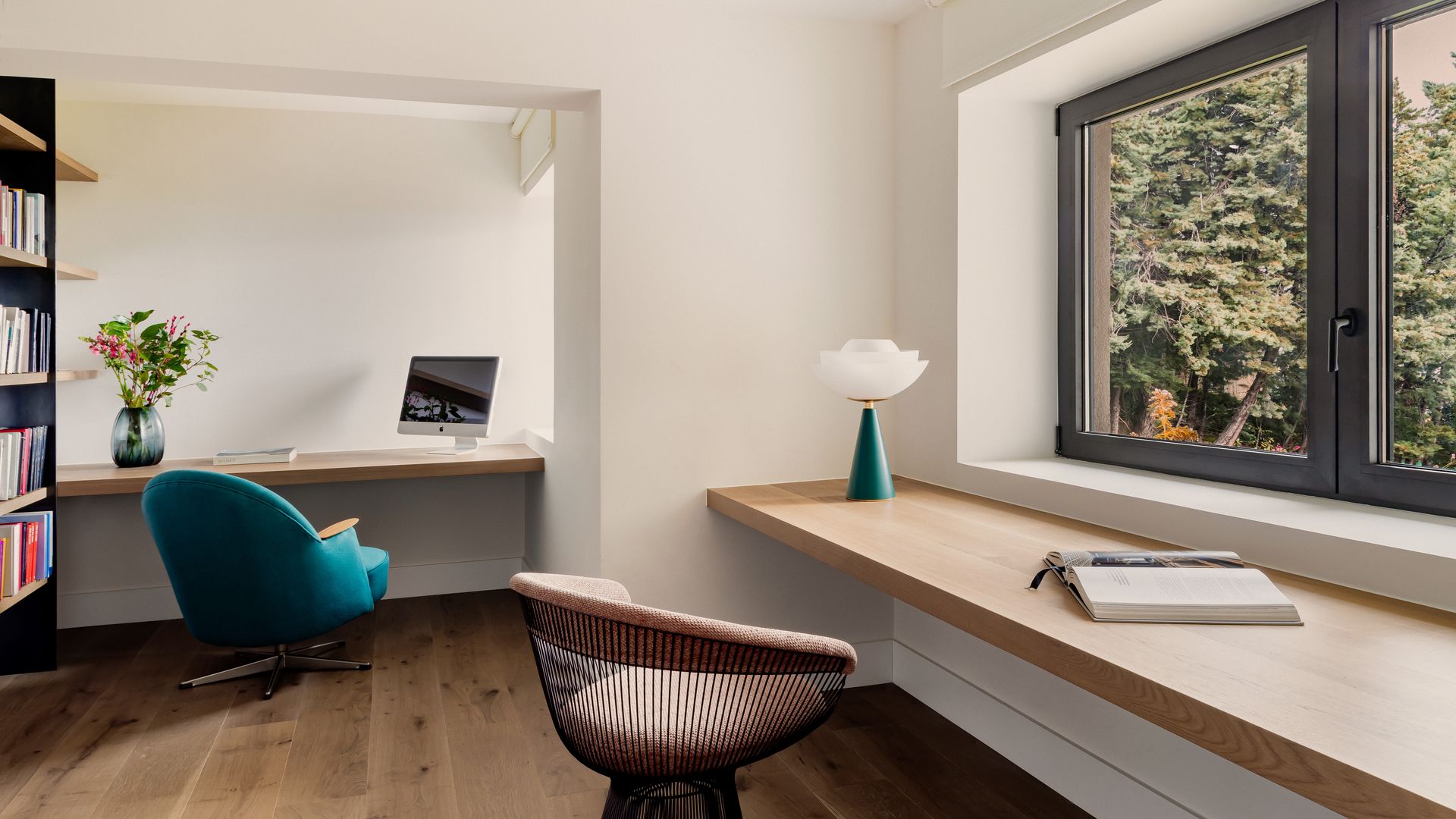 Despacho con dos mesas bajo las ventanas colocadas en L, sillas, estantería negra, lámpara de sobremesa, jarrón 