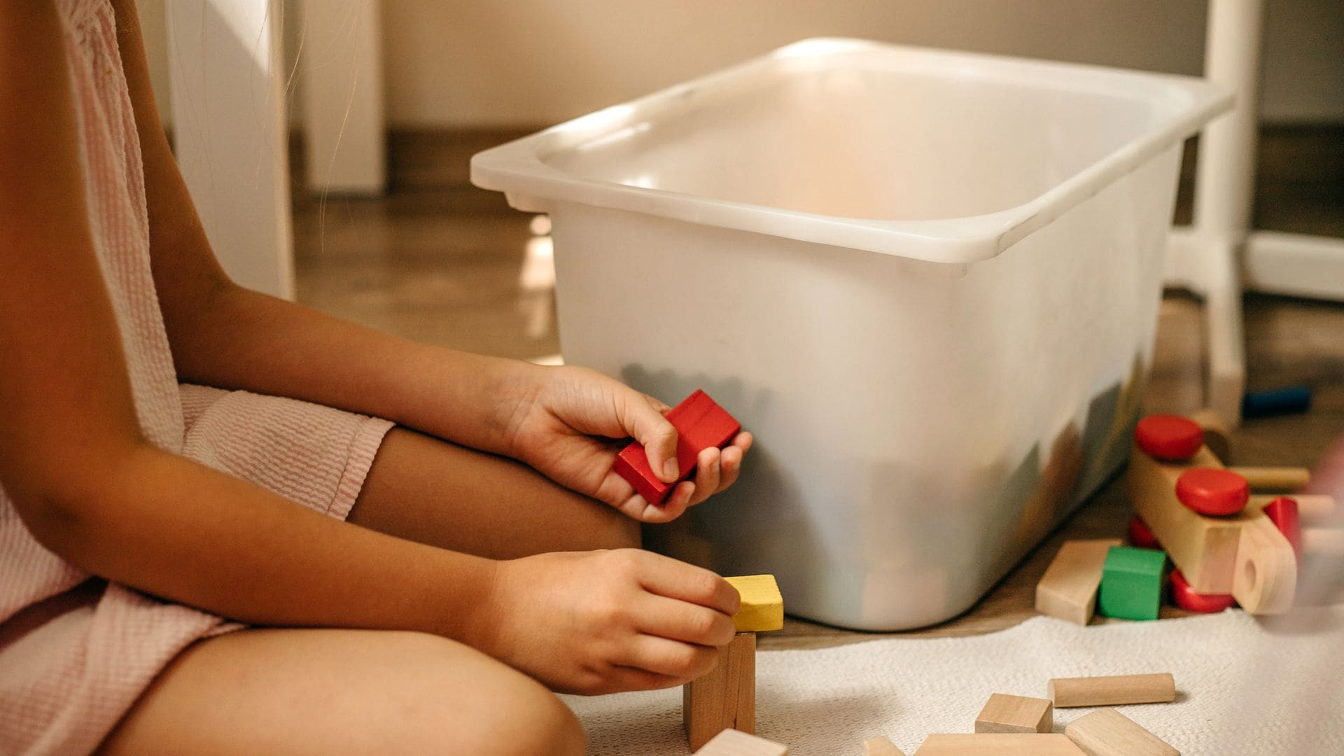 Niña pequeña con un juego de construcciones de madera en el suelo, con una caja TROFAST de IKEA detrás.