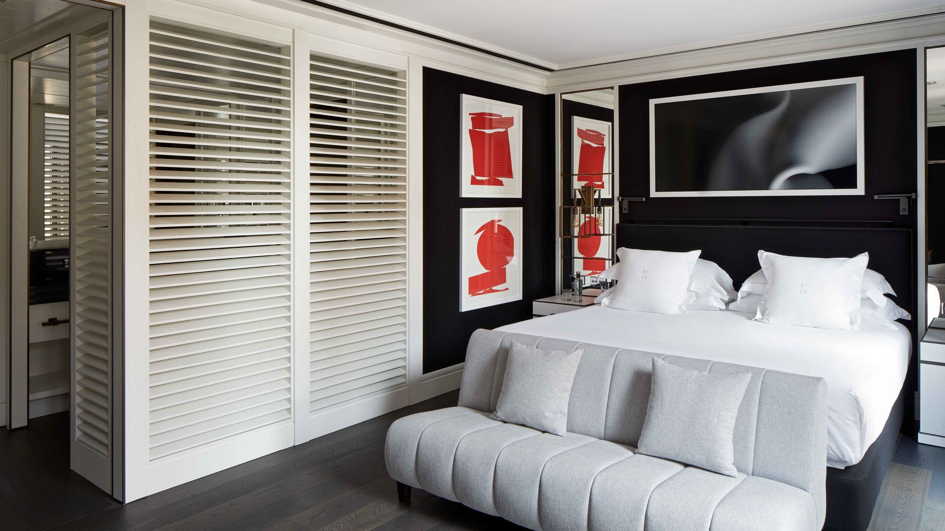 Dormitorio con baño integrado, pared cabecero negra, baño tras listones de madera blanco, cuadros rojos en la pared, banco con cojines 