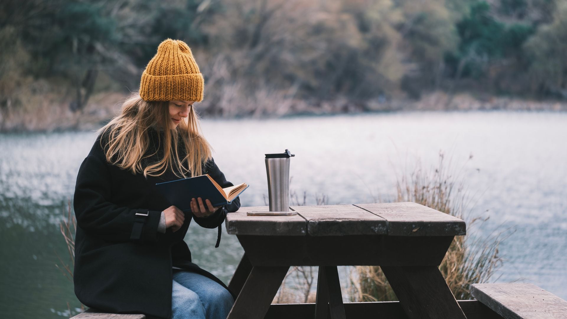 Mujer leyendo un libro en un lago, sentada en una mesa de picnic de madera, con ropa de invierno y gorro. Tiene un a bebida caliente en un termo sobre la mesa.