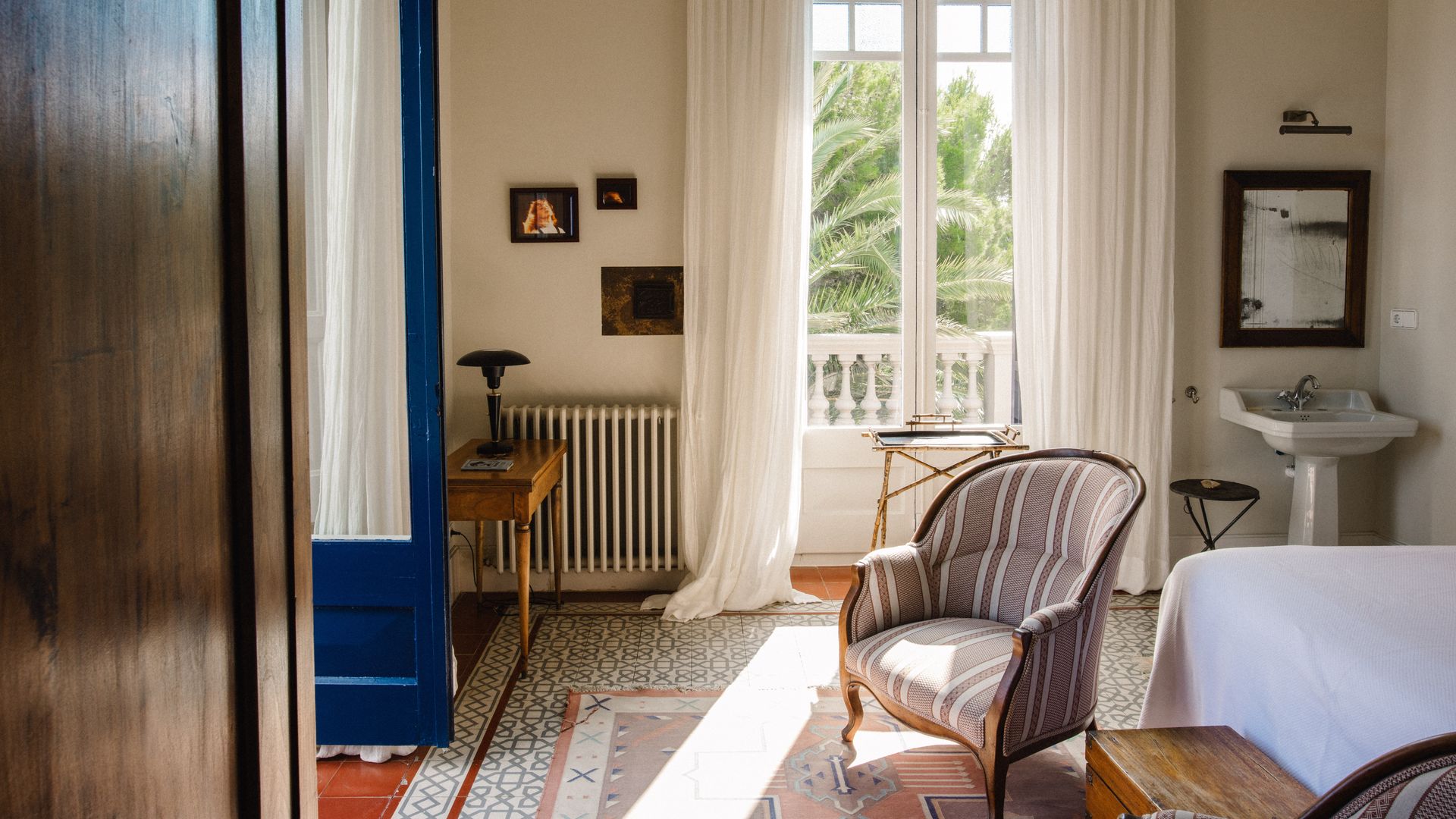 Dormitorio con salida a la terraza con puerta azul, lavabo de pedestal incluido en la habitación, butaca, suelo de barro, cortinas blancas