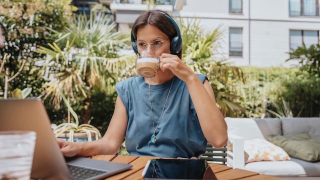 Mujer teletrabajando en una terraza con sol, con un ordenador y sus auriculares, además de estar bebiendo un café.