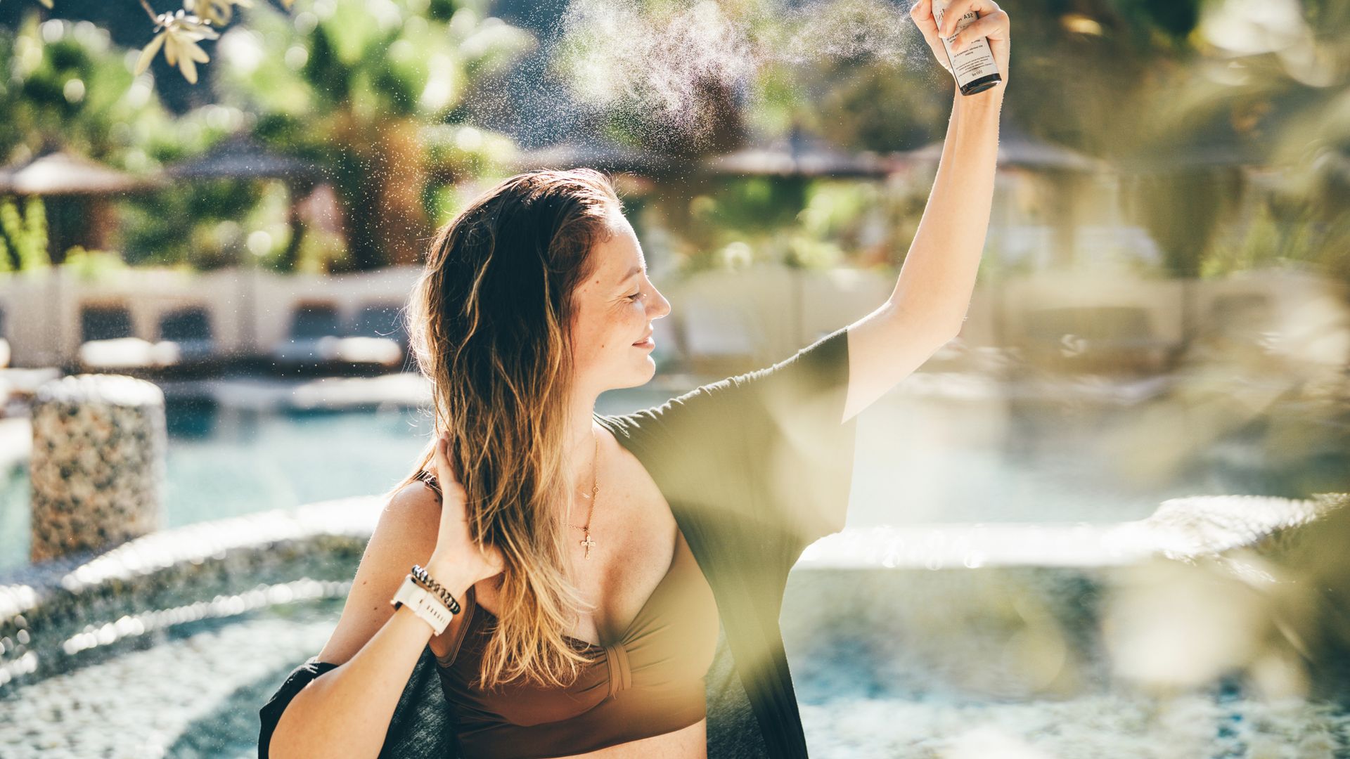 Mujer con pelo castaño claro sentada en una piscina aplicándose spray en el cabello 