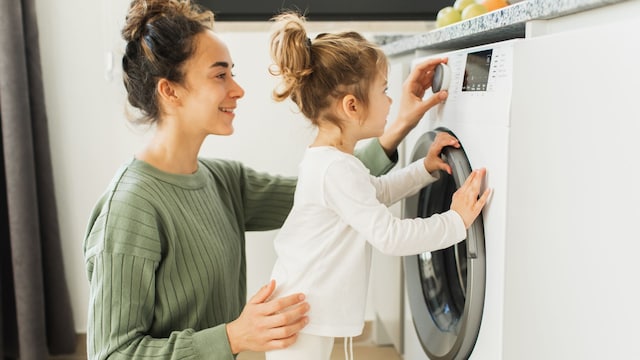 Madre poniendo con su hija la lavadora en su casa, ambas sonriendo y colaborando juntas.