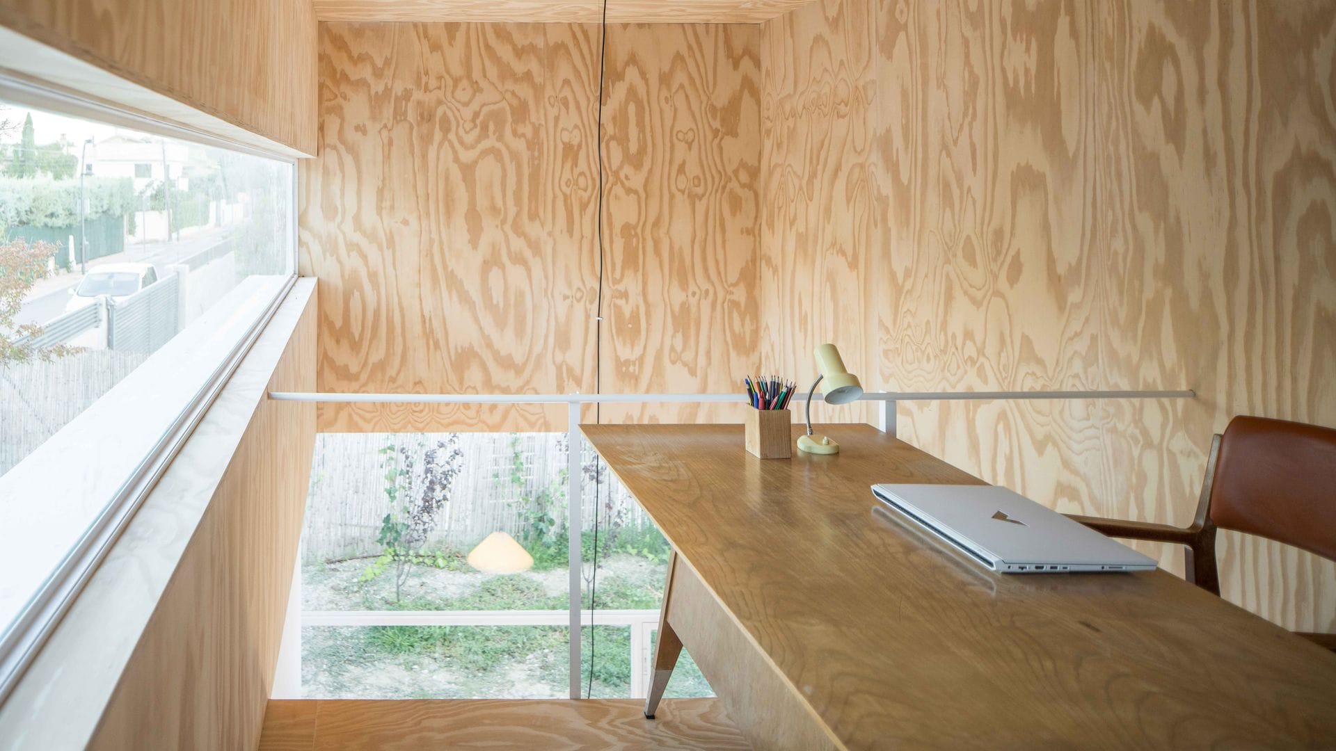 Despacho con paredes, mesa y suelo de madera, silla de cuero, ventanal y flexo amarillo