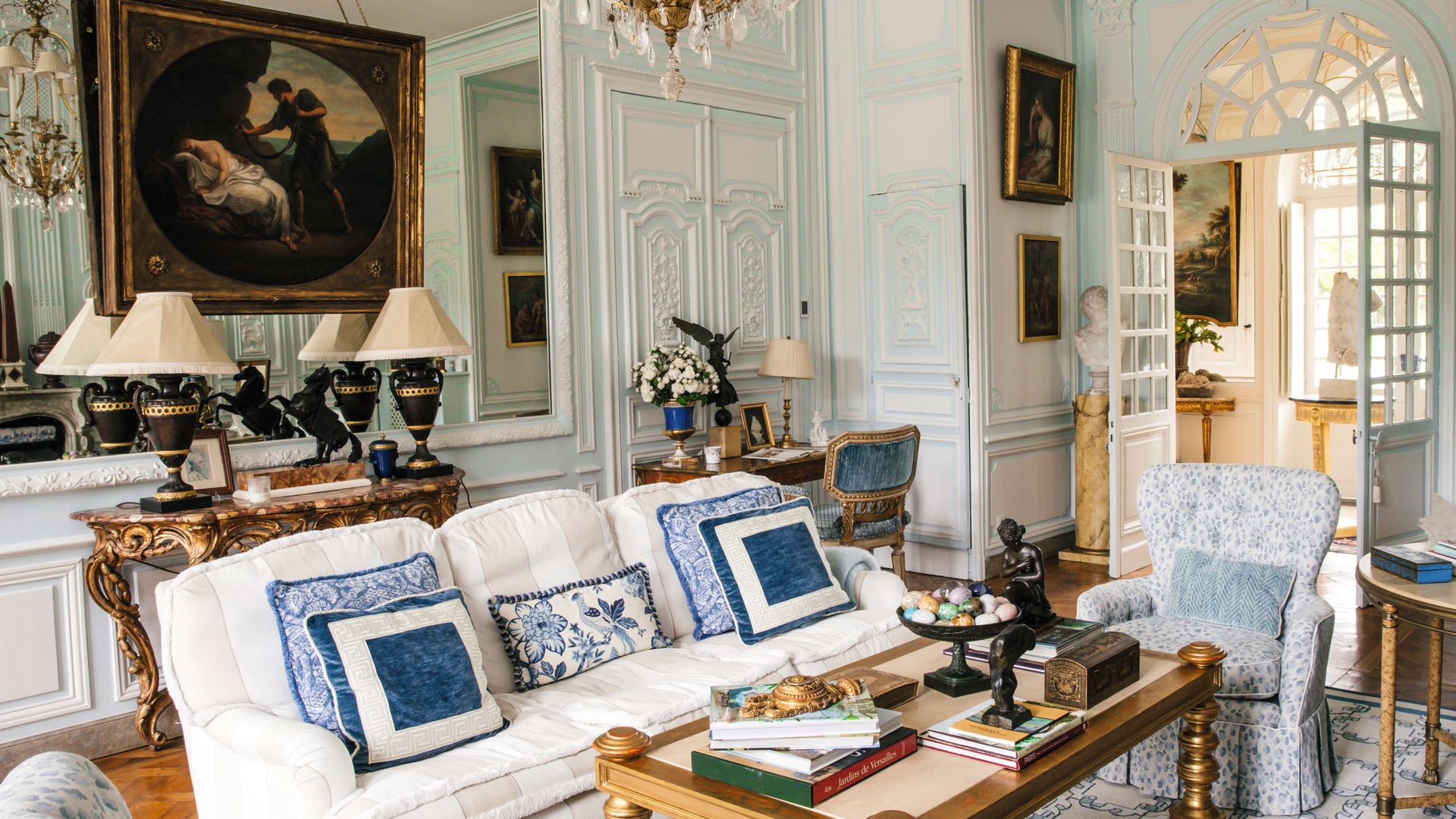 Sala de estar elegante con sofá blanco, cojines azules, mesa de café con libros y una pintura grande en la pared.