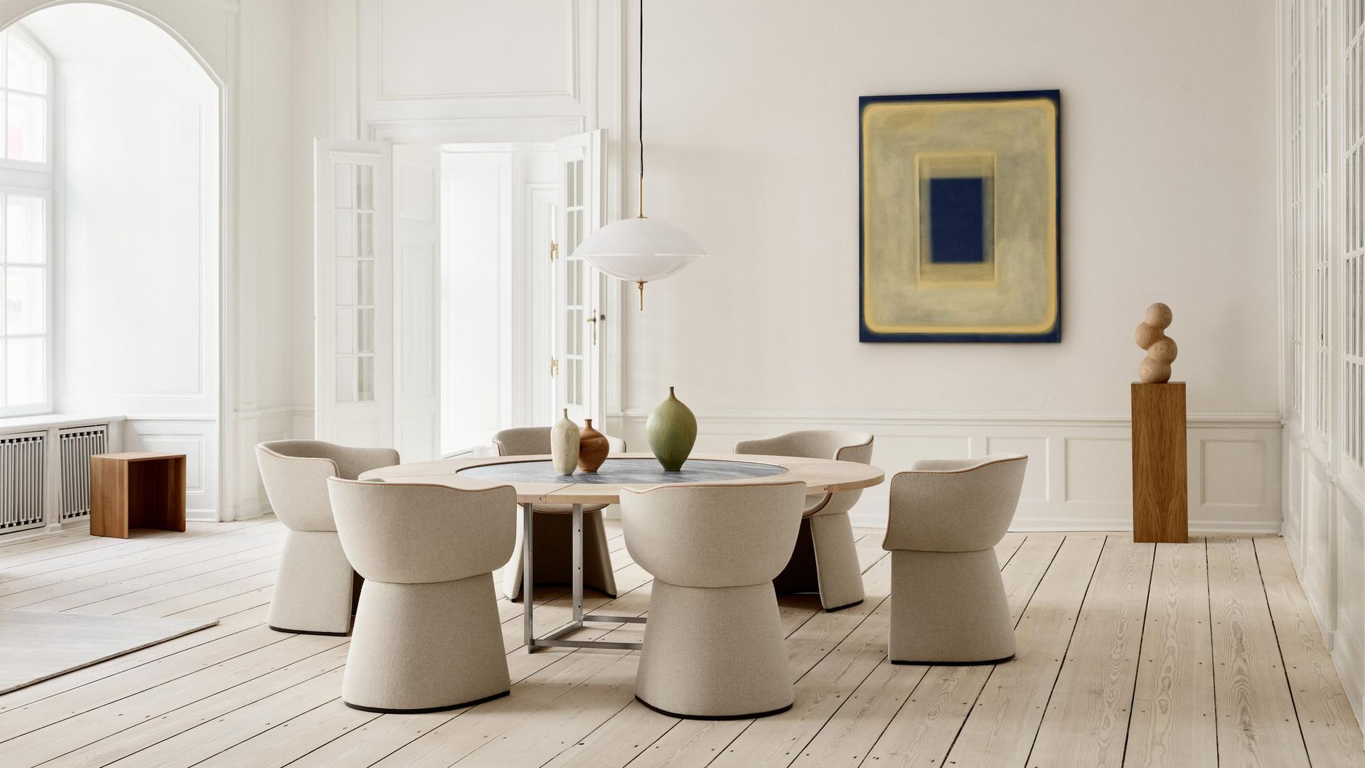 Comedor con suelo de madera blanco, mesa redonda, sillas tapizadas con forma curva, lámpara de techo, molduras, cuadro en la pared, pedestal madera