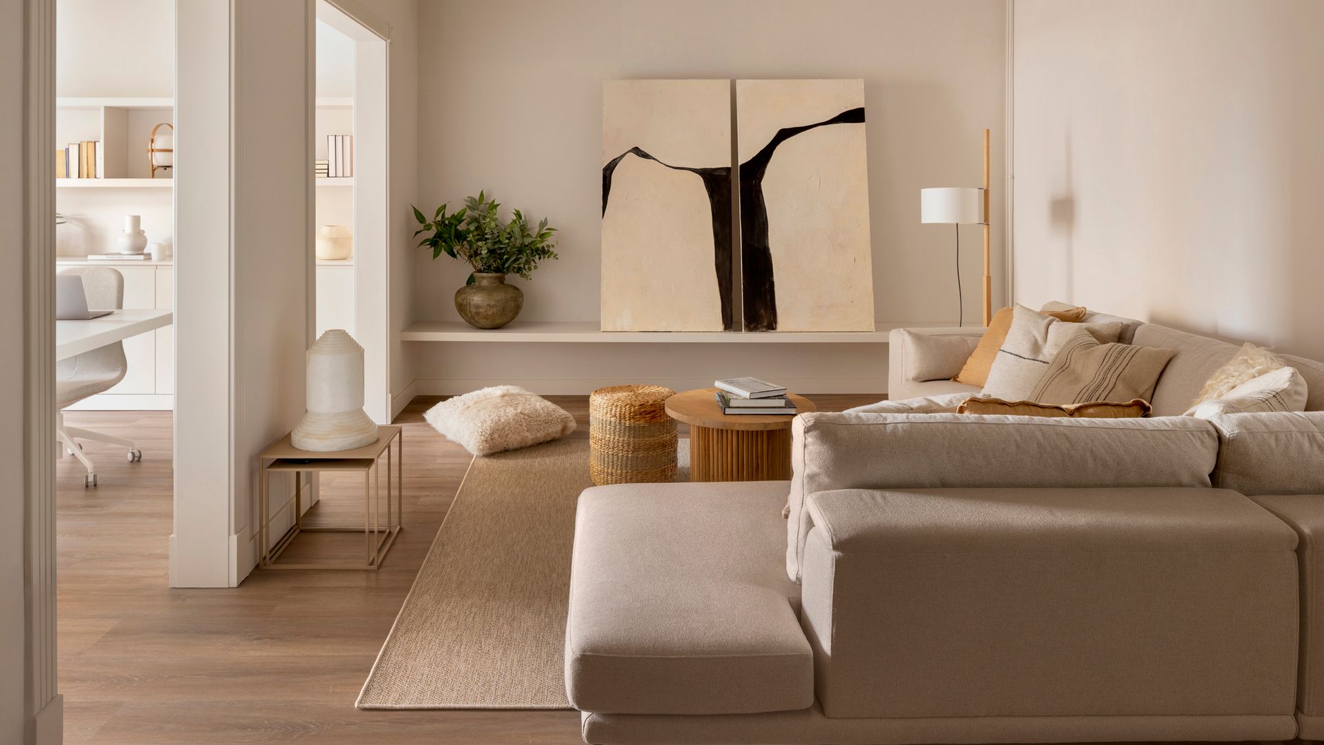 Salón con sofá beige, comunicado con despacho. balda en la pared, cuadRo apoyado sobre la balda, alfombra, mesa de centro