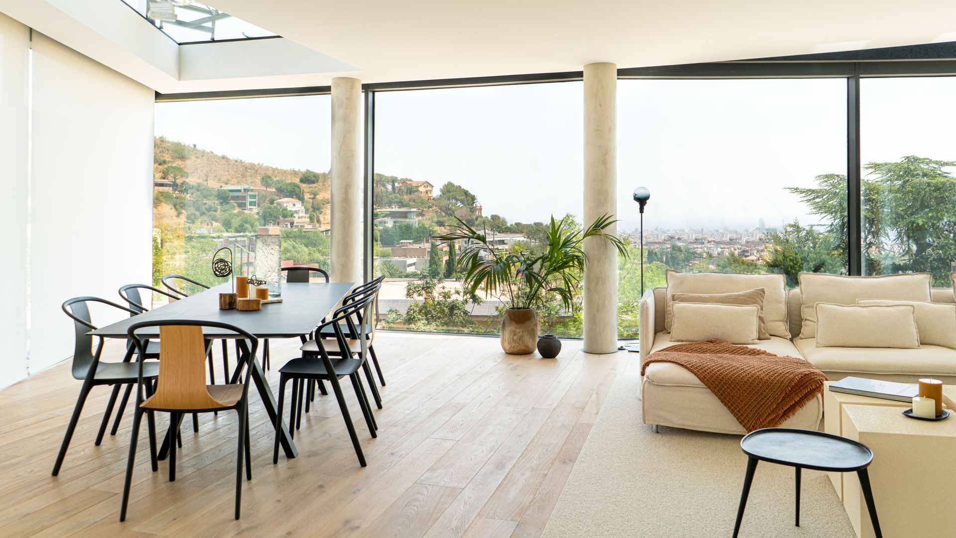 Salón comedor con grandes ventanales con vistas jardín, columnas, sofás blancos, suelo de madera claro, mesa negra y sillas a juego, alfombra blanca