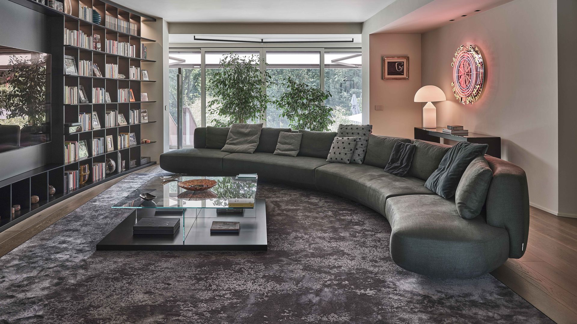 Salón con sofá gris curvo, grandes cristaleras con vistas al jardín, librería negra de suelo a techo, alfombra gris, mesa de centro con el sobre de cristal