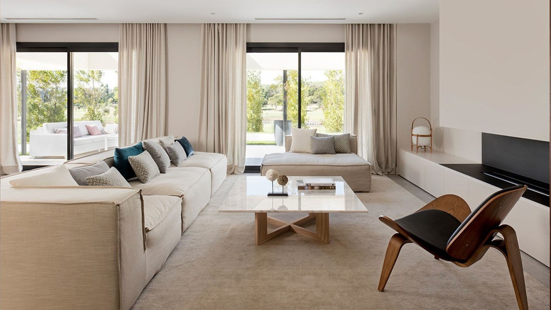 Salón en tonos beiges con salida al jardín, grandes ventanales, cortinas blancas. sofá blanco roto, alfombra, mesa de centro de cristal, mueble bajo de televisión, lámpara Cesta