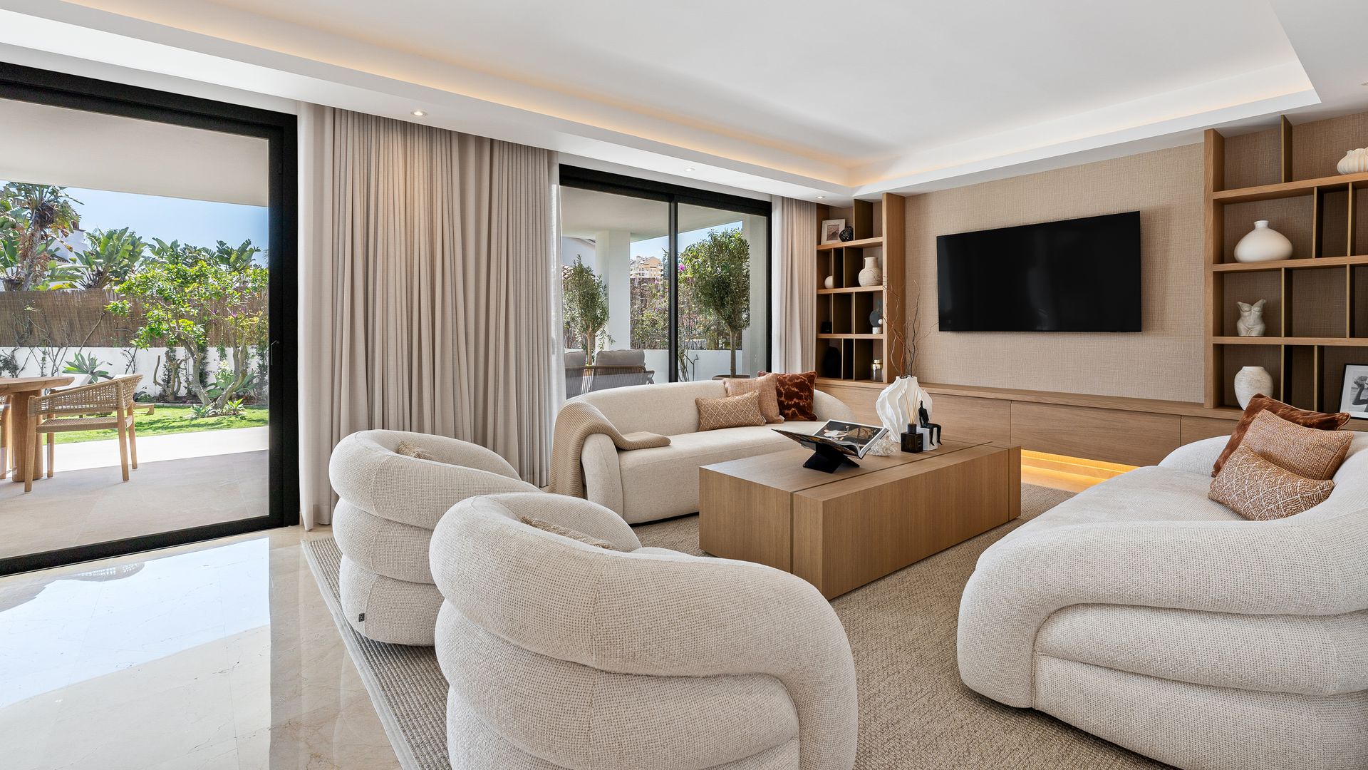Salón abierto al jardín con mesa de centro de madera, alfombra, sofás y butacas blancos, televisión en la pared, estructura de madera con estantería, cortinas blancas