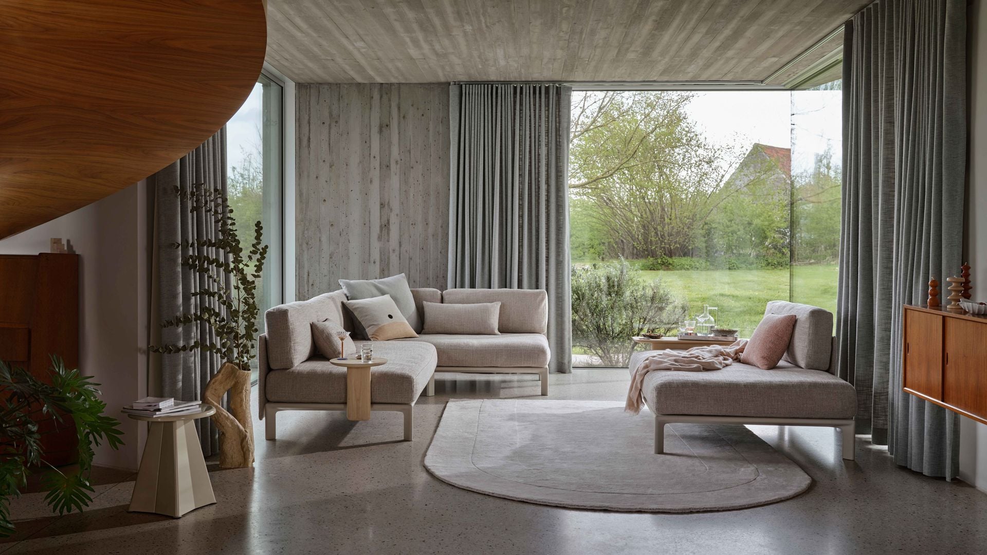 Salón con gran ventanal, suelo de cemento, sofá gris, alfombra y aparador de madera