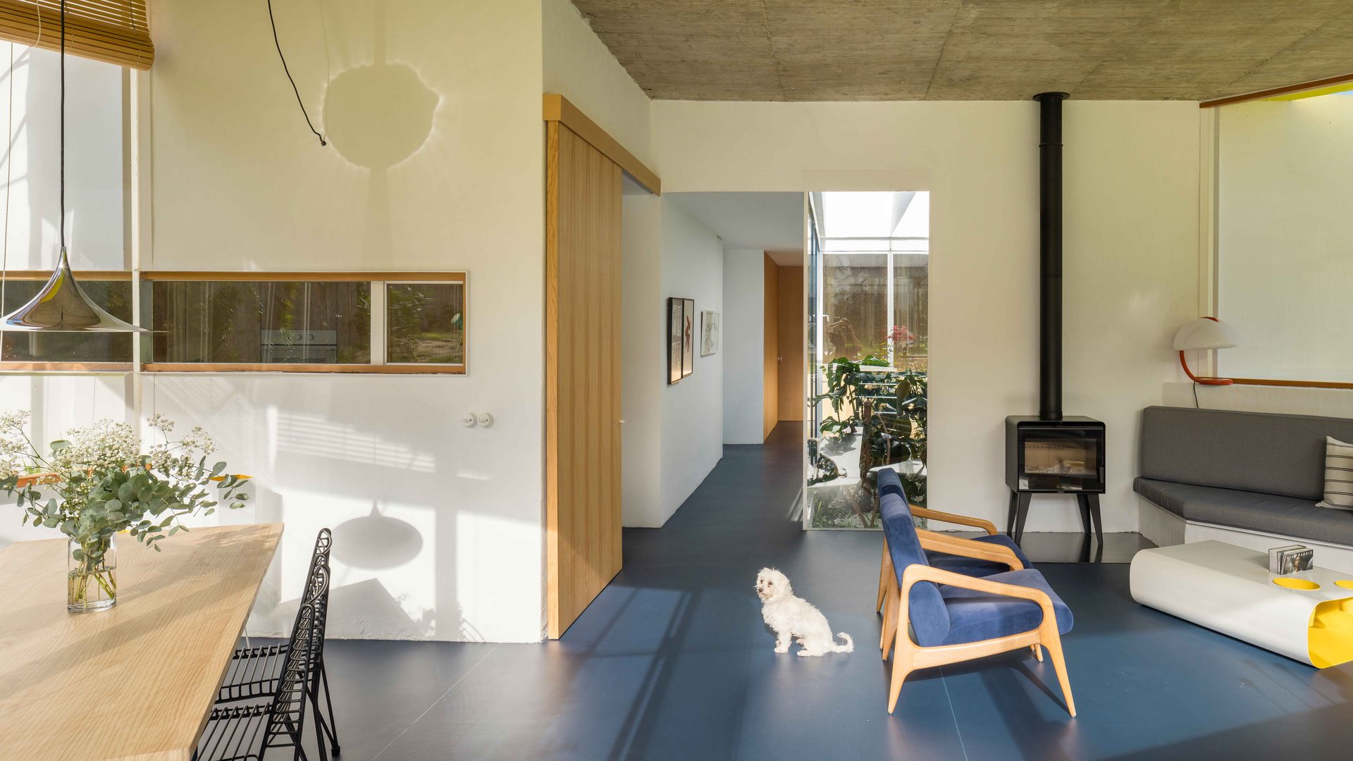 Salón comedor grandes ventanales, banco de obra con cojines grises, suelo azul, mesa de madera y sillas metálicas