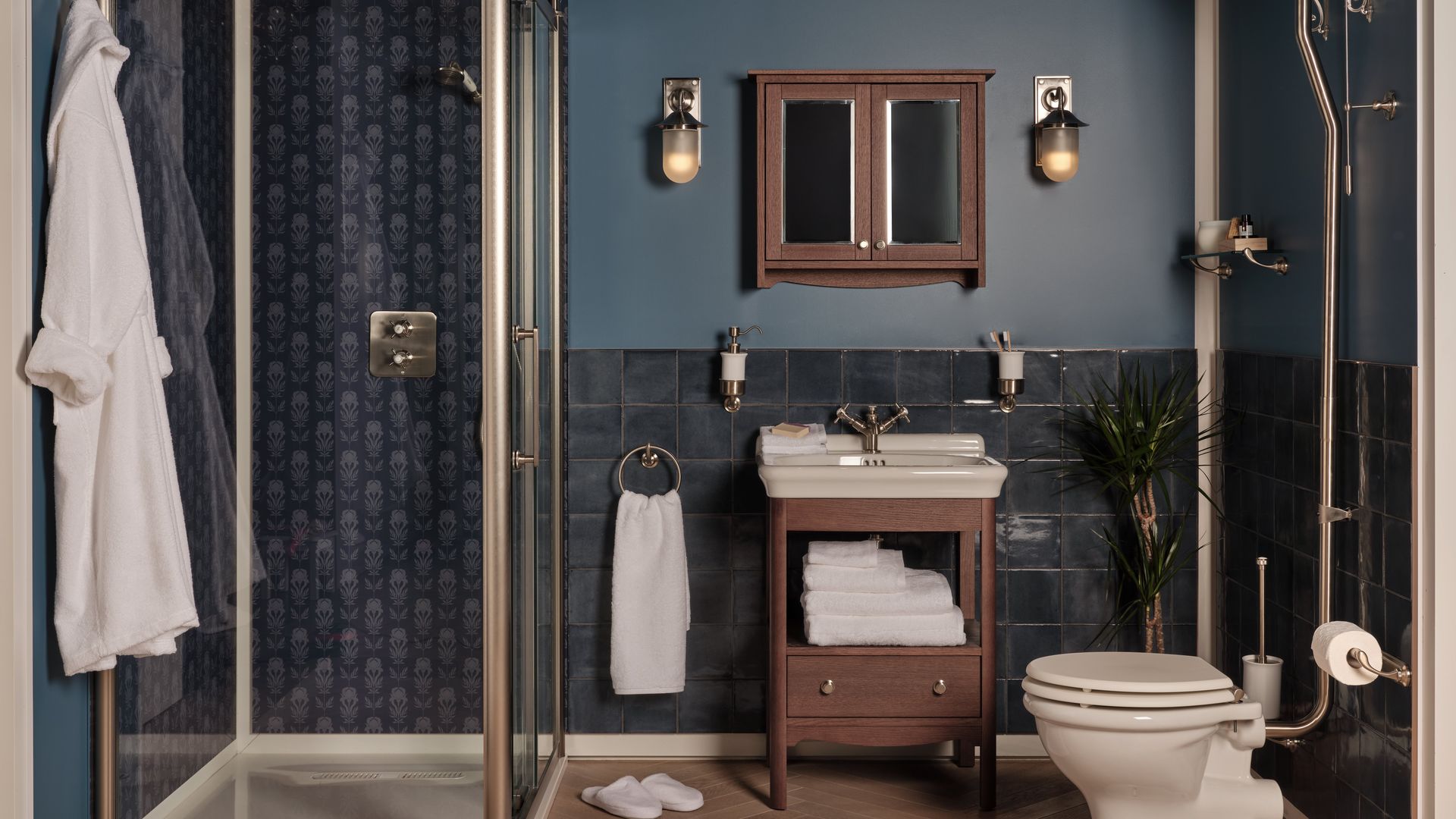 Baño en color azul con ducha y mampara dorada, toallas blancas, mueble bajolavabo de madera, apliques de pared, espejo con armario