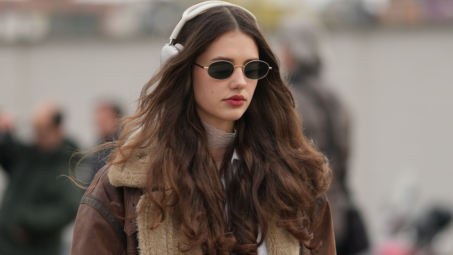 Mujer joven con pelo castaño largo y ondulado, cazadora de pelo y gafas de sol en Milan