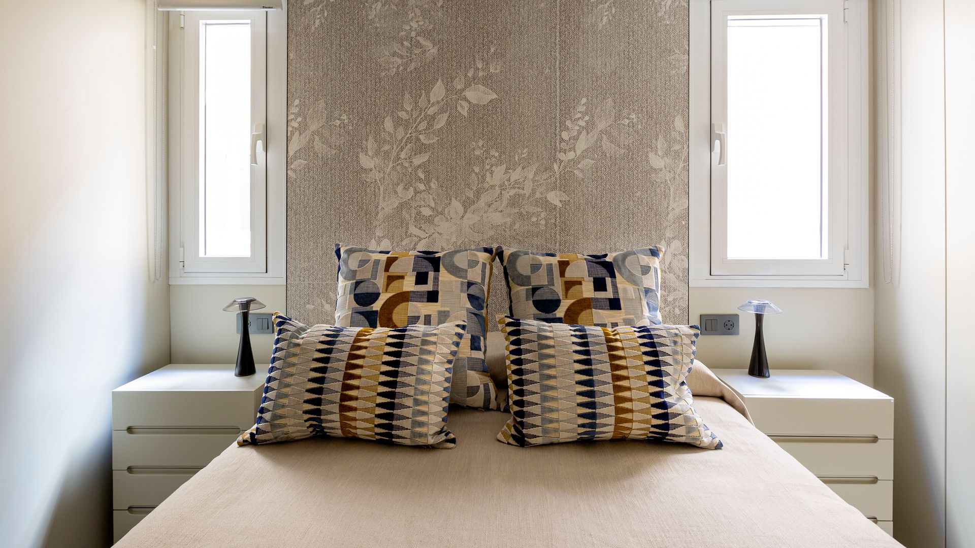 Dormitorio con pared del cabecero de papel pintado, cojines sobre la cama, mesitas de noche de color blanco, ventanas a ambos lados de la cama