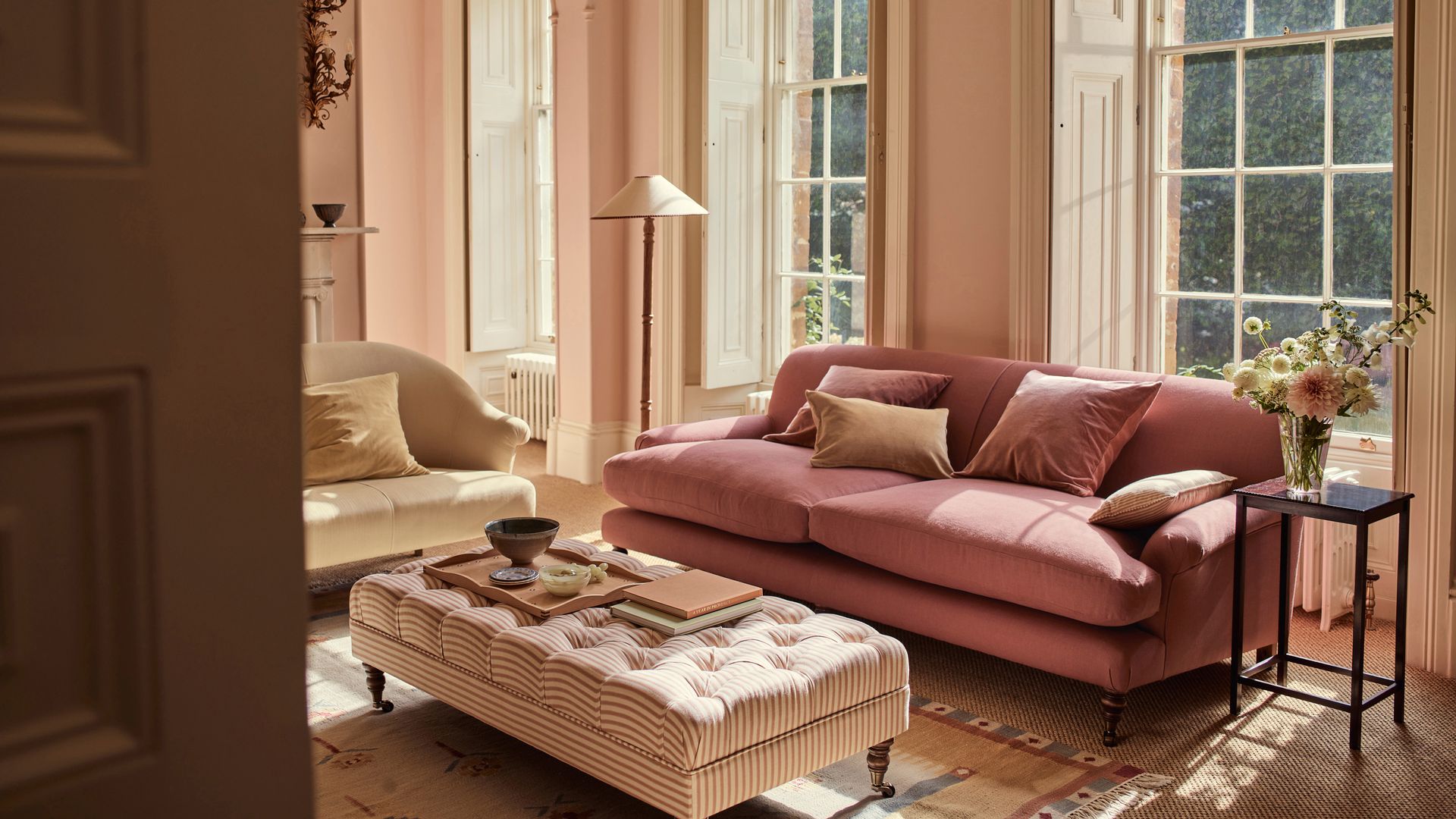Salón clásico con mesa tapizada de capitoné, sofá rosa, sillón beige, grandes ventanales