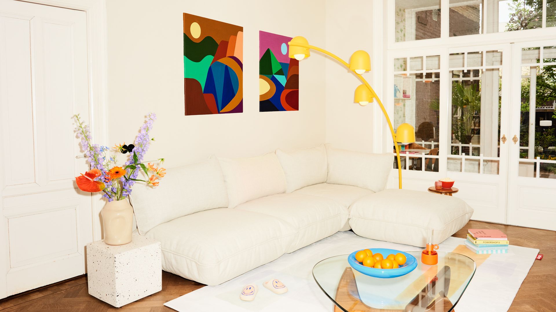Salón con sofá blanco, alfombra blanco, puf, mesa de centro de cristal, lámpara amarilla de pie, cuadros en la pared