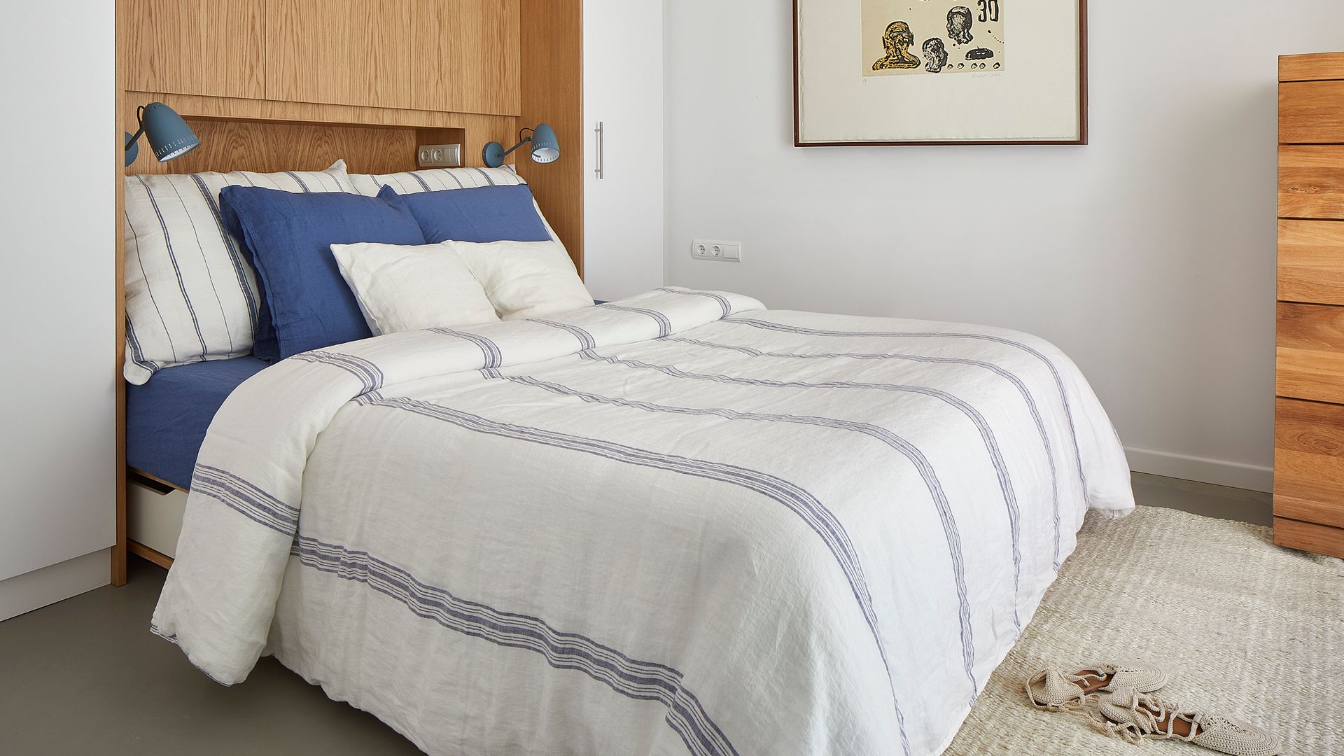Dormitorio con estructura a medida sobre la pared del cabecero con armarios en lo alto del techo, cabecero de madera con balda, ropa de cama de rayas blancas y azules