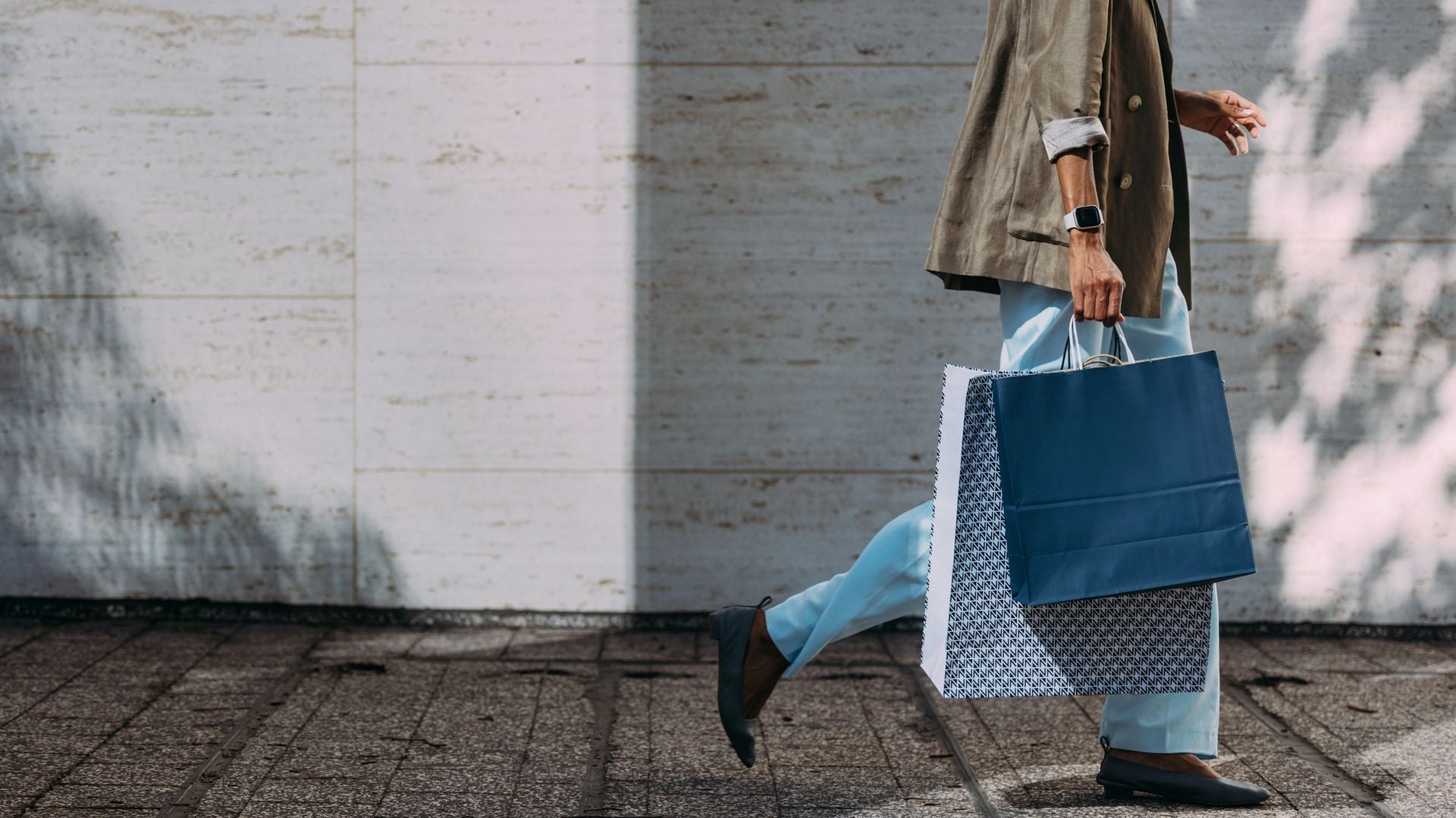 Mujer caminando con bolsas de compra por la calle, con ropa de otoño (pantalones vaqueros y blazer camel). Solo se le ve hasta la altura del codo.